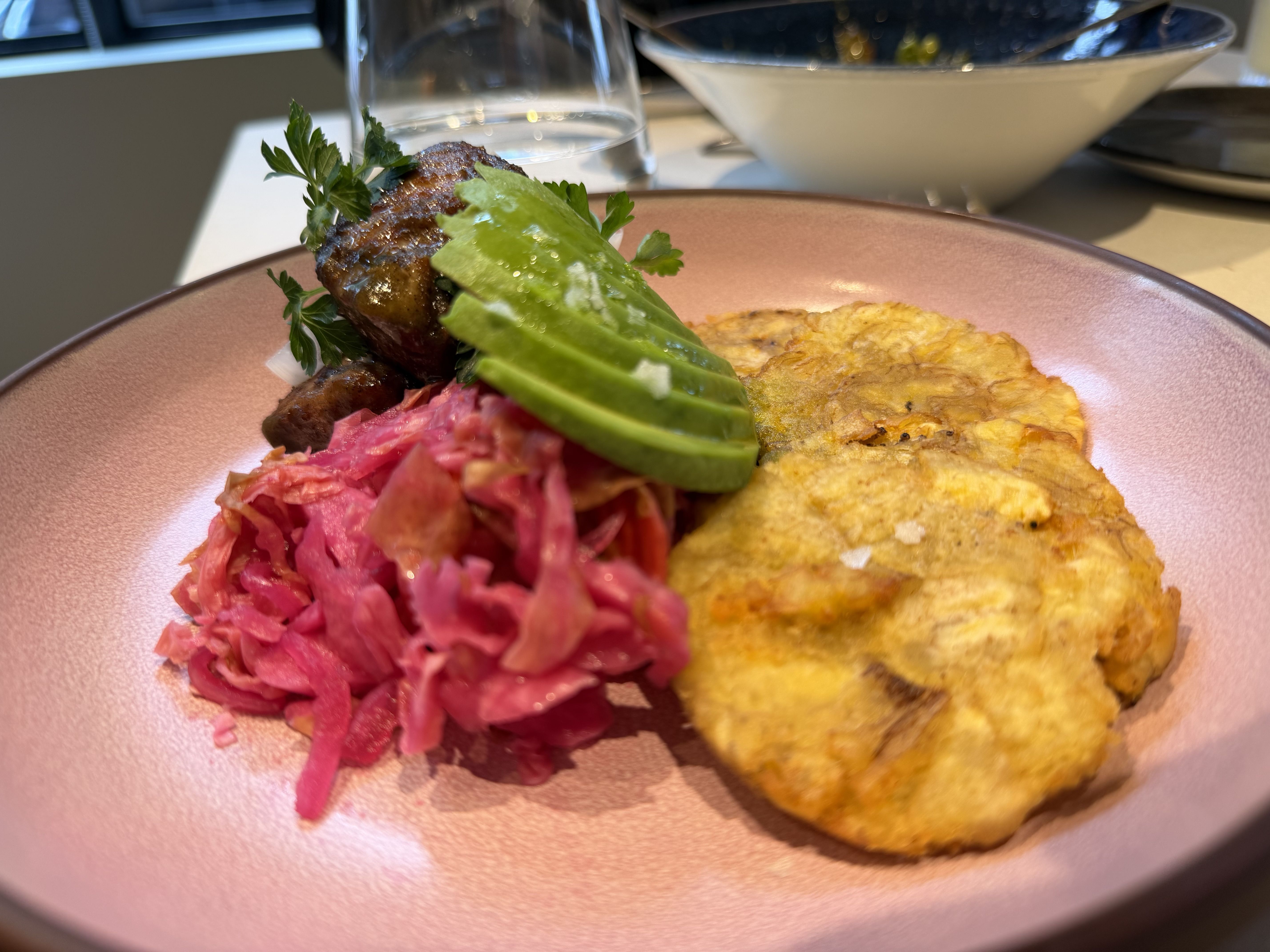 Close-up of a pink plate featuring bright pink shredded cabbage, a golden potato pancake, and a herb-garnished meat piece with parsley. In the background, a glass of water and a white bowl.