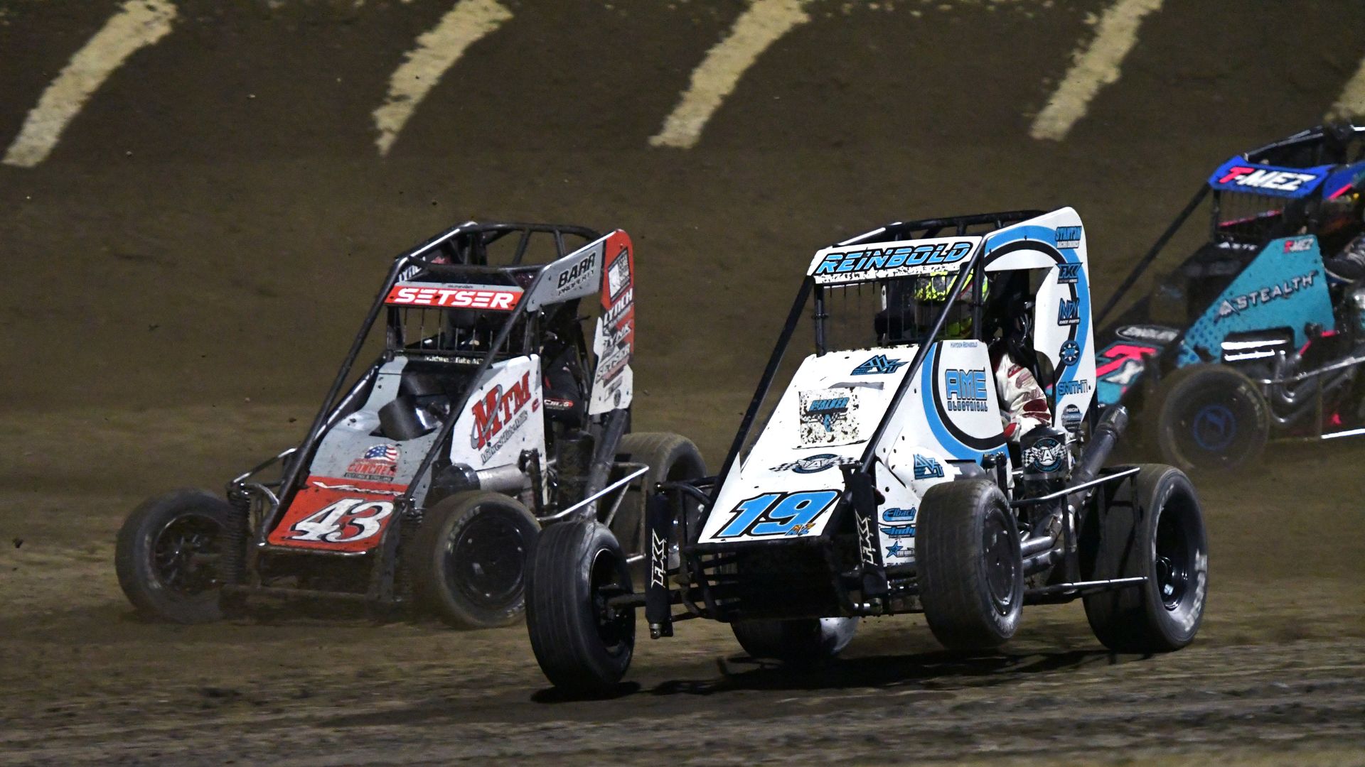 Three dirt track sprint cars racing on a dirt oval at night; car 43 in orange and black, car 19 in white and blue, and a third car mostly blue in the background.