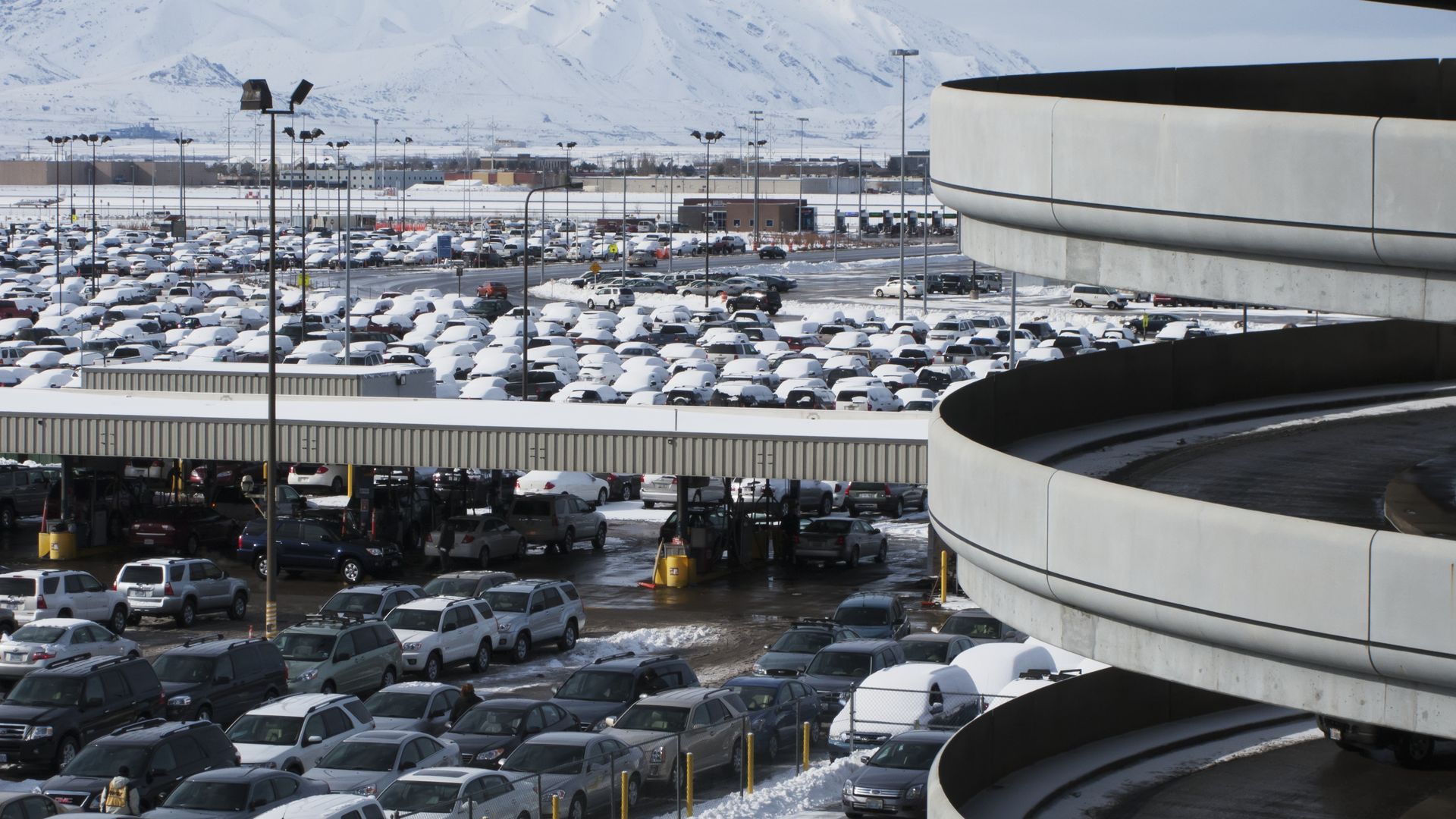 Snow-covered parking lot full of cars, with a large circular concrete parking ramp on the right. Snowy mountains rise in the distance under a pale blue sky.