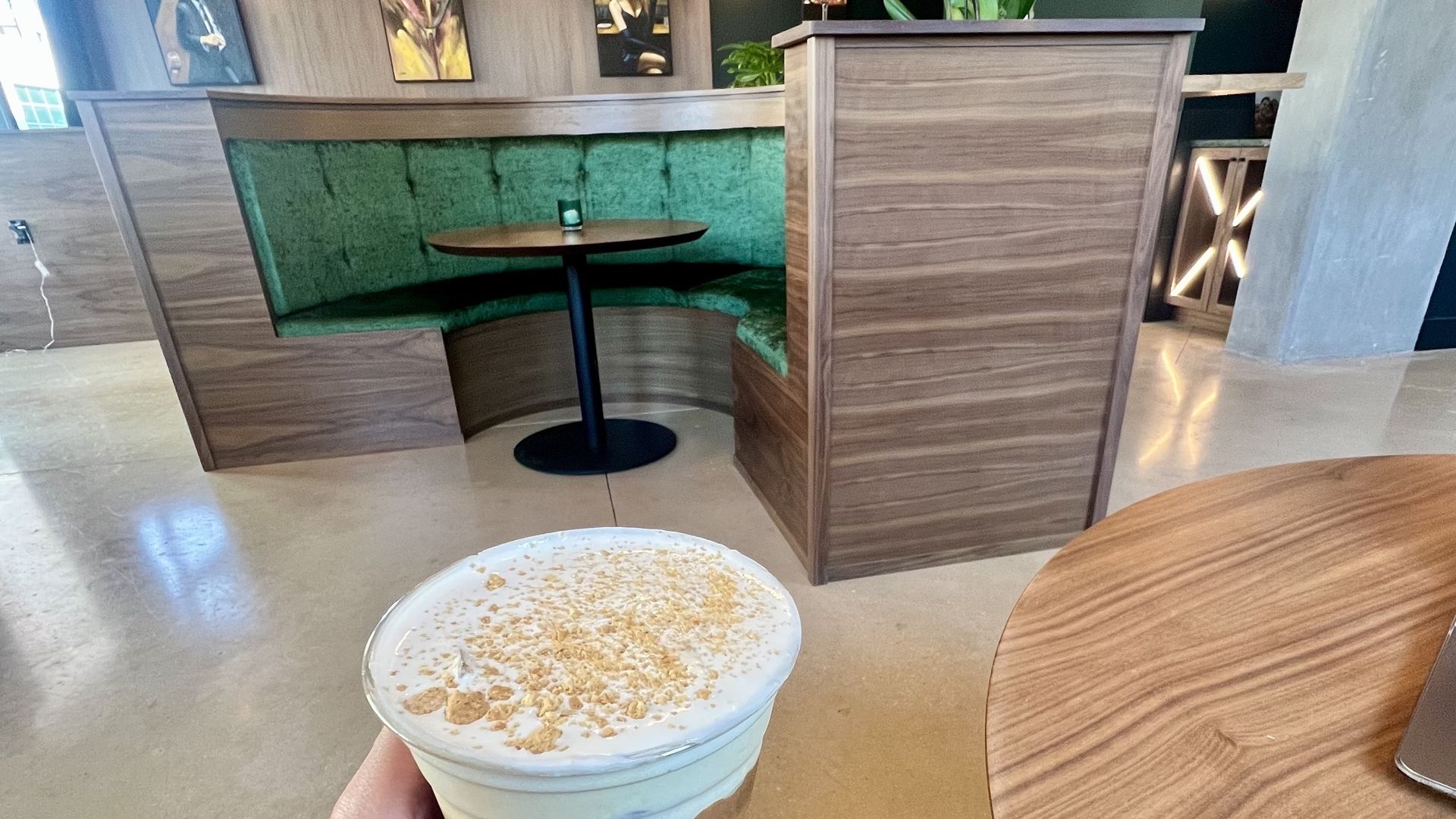 A hand holding an iced latte topped with cream and crumbled cookie topping inside a café with green velvet booth seating, wooden tables and framed art on the walls. The photo shows the caramel cookie butter iced latte at Kafe Krave in San Antonio.