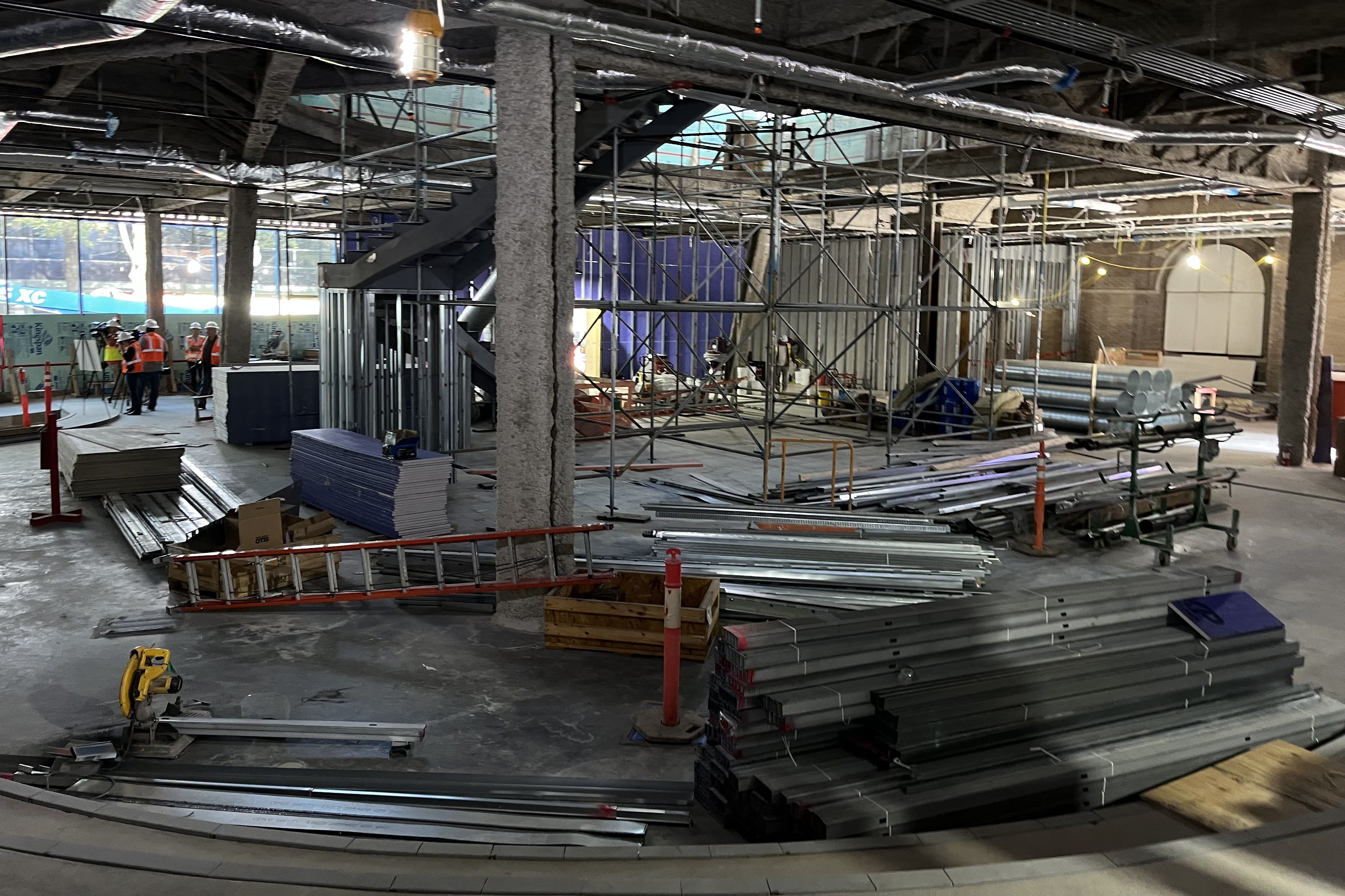 Indoor construction site with steel beams, scaffolding, stacks of drywall, a red ladder, and workers in orange safety vests and helmets near a large window letting in daylight.