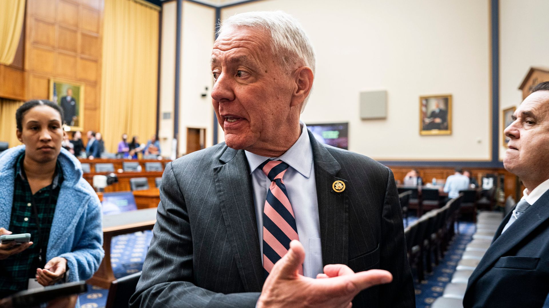 Rep. Ken Buck, wearing a gray suit, light blue shirt and blue and pink striped tie, points off to the side in a congressional hearing room.