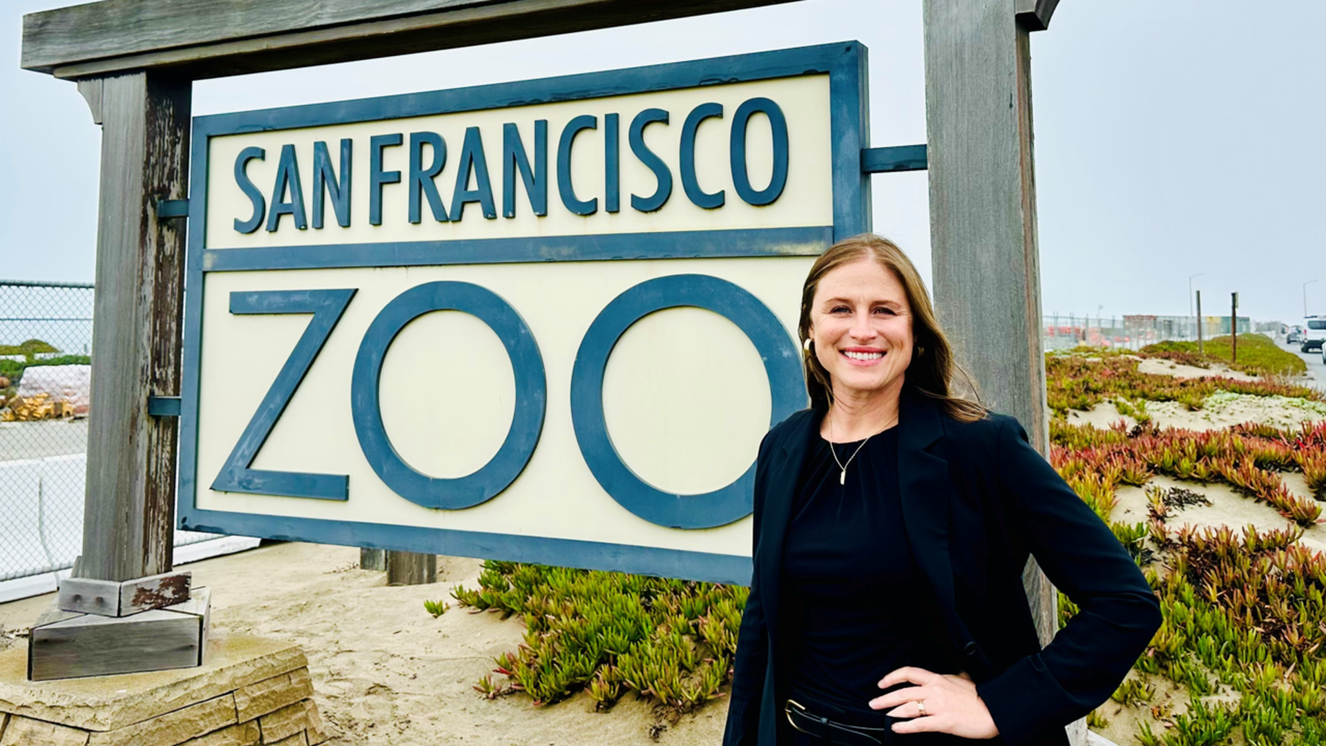 Smiling woman in black outfit stands with hand on hip next to large San Francisco Zoo sign featuring blue animal silhouettes atop wooden frame on sandy ground.