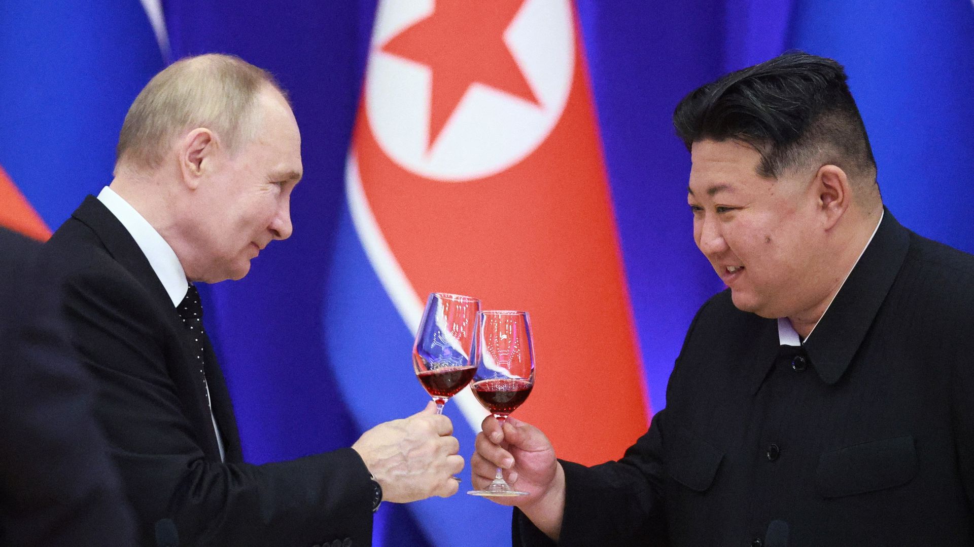 Vladimir Putin and Kim Jong Un, both dressed in dark formal wear, clink glasses full of red wine. 