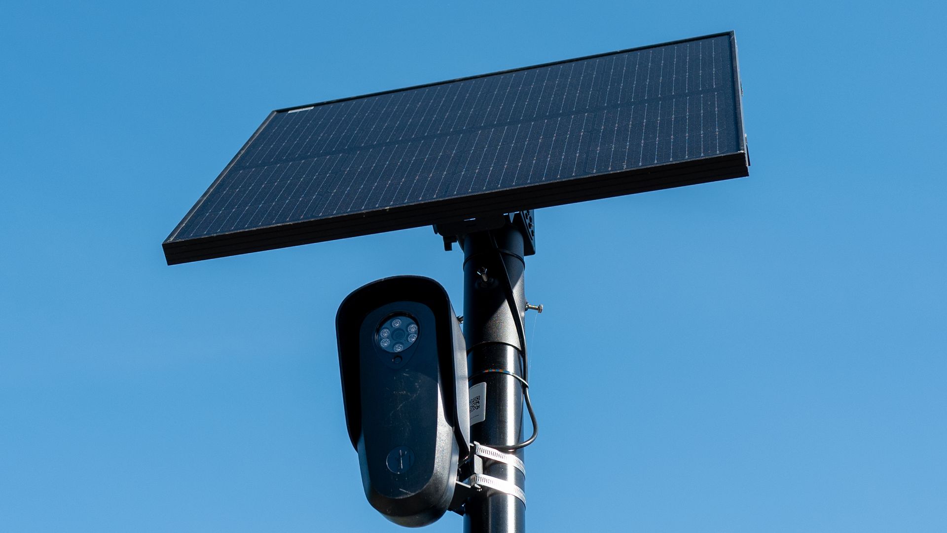 Tilted solar panel mounted on a pole above a traffic signal, with a round LED light housing beneath, set against a clear blue sky.