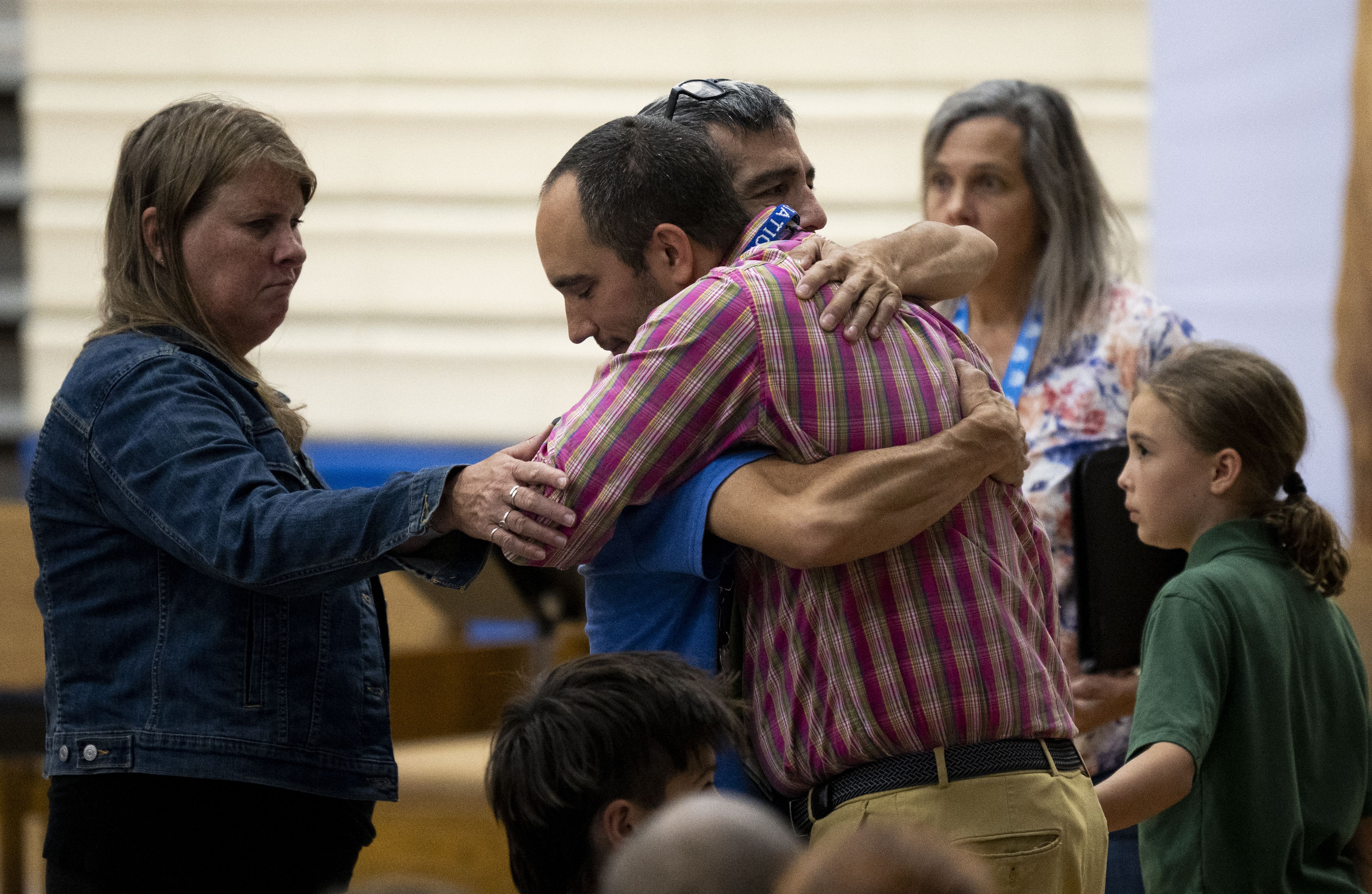 A group hug indoors: a man in a pink plaid shirt hugs a man in blue, with a woman in a denim jacket on the left and others nearby watching.