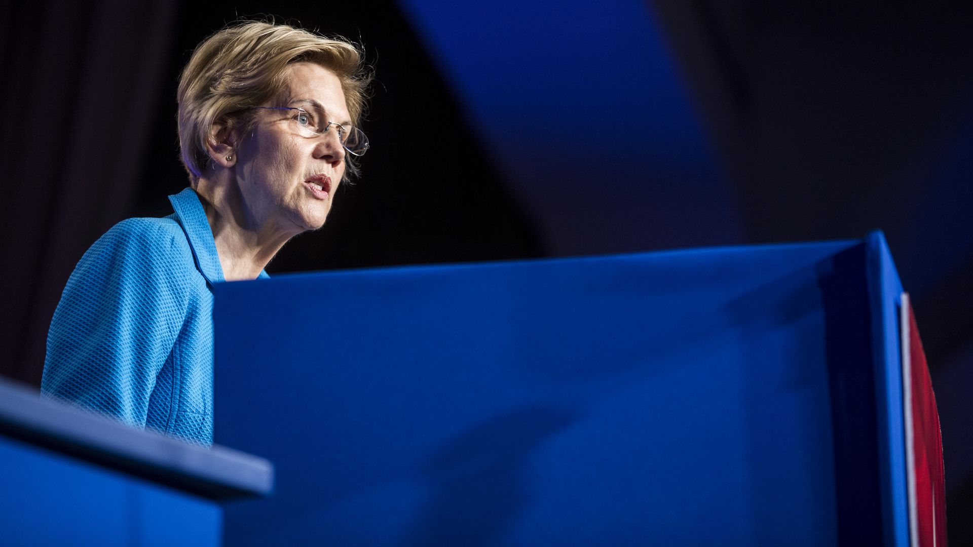 In this image, Elizabeth Warren stands and speaks behind a blue podium.
