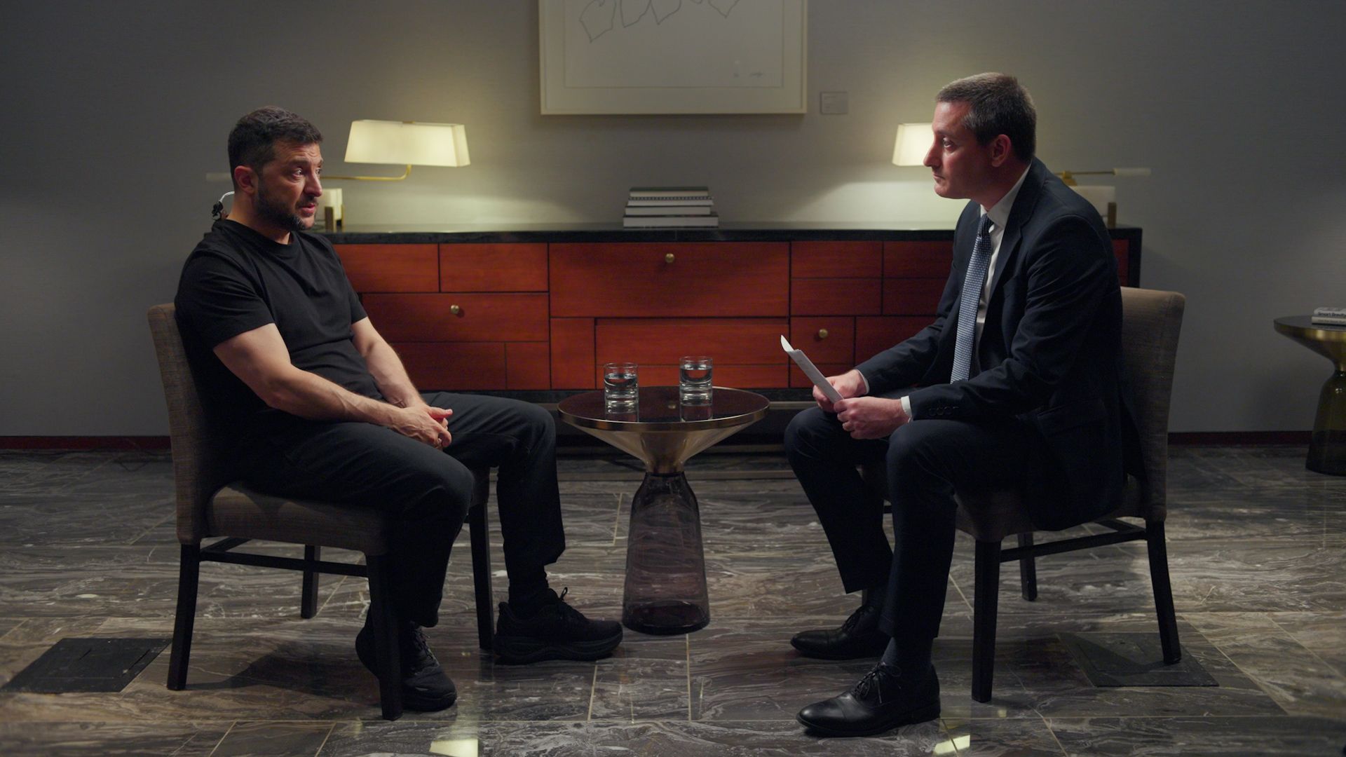 Volodymyr Zelensky and Axios' Barak Ravid sit facing each other in a formal interview setting; Zelensky in a black t-shirt, Barak in a dark suit, with a small table holding two glasses of water between them.