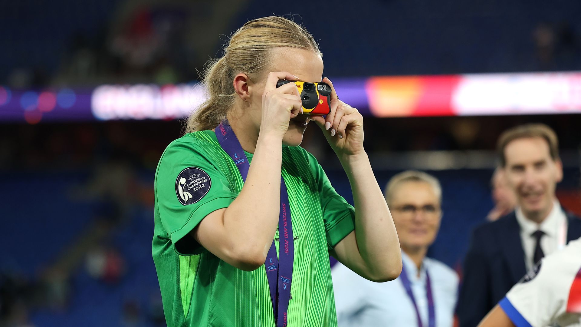 Person in green sports jersey with UEFA Euro 2022 patch and gold medal using a yellow and red disposable camera, with blurred people and stadium lights in the background.