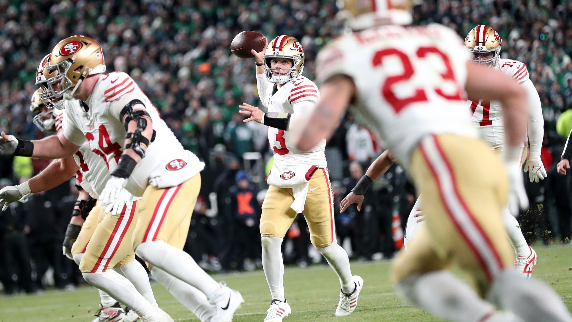 San Francisco 49ers quarterback prepares to throw a football with teammates in white and gold uniforms and gold helmets on a green field during a night game.