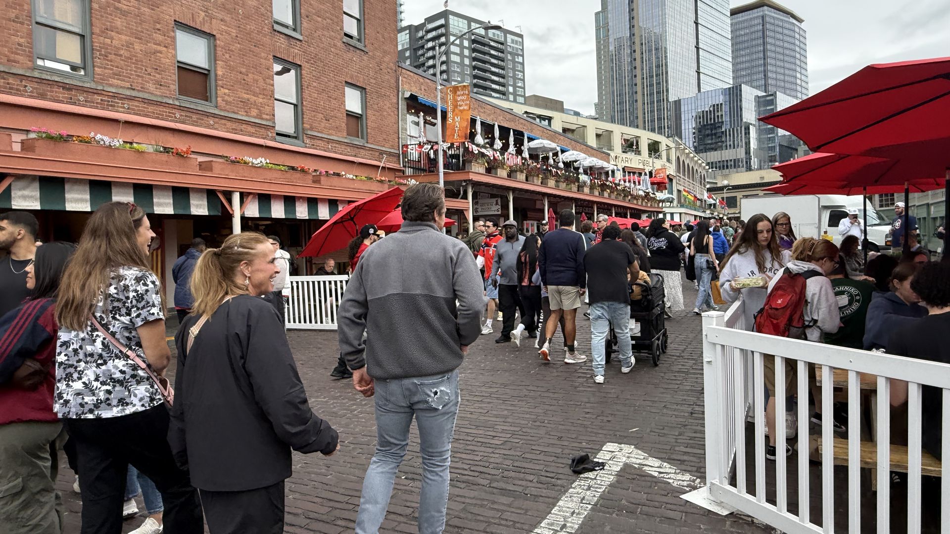 Busy urban street market with red brick row houses, colorful flower boxes, and red umbrellas lining stalls. A crowd walks on cobblestone, with tall glass skyscrapers in a cloudy sky behind.
