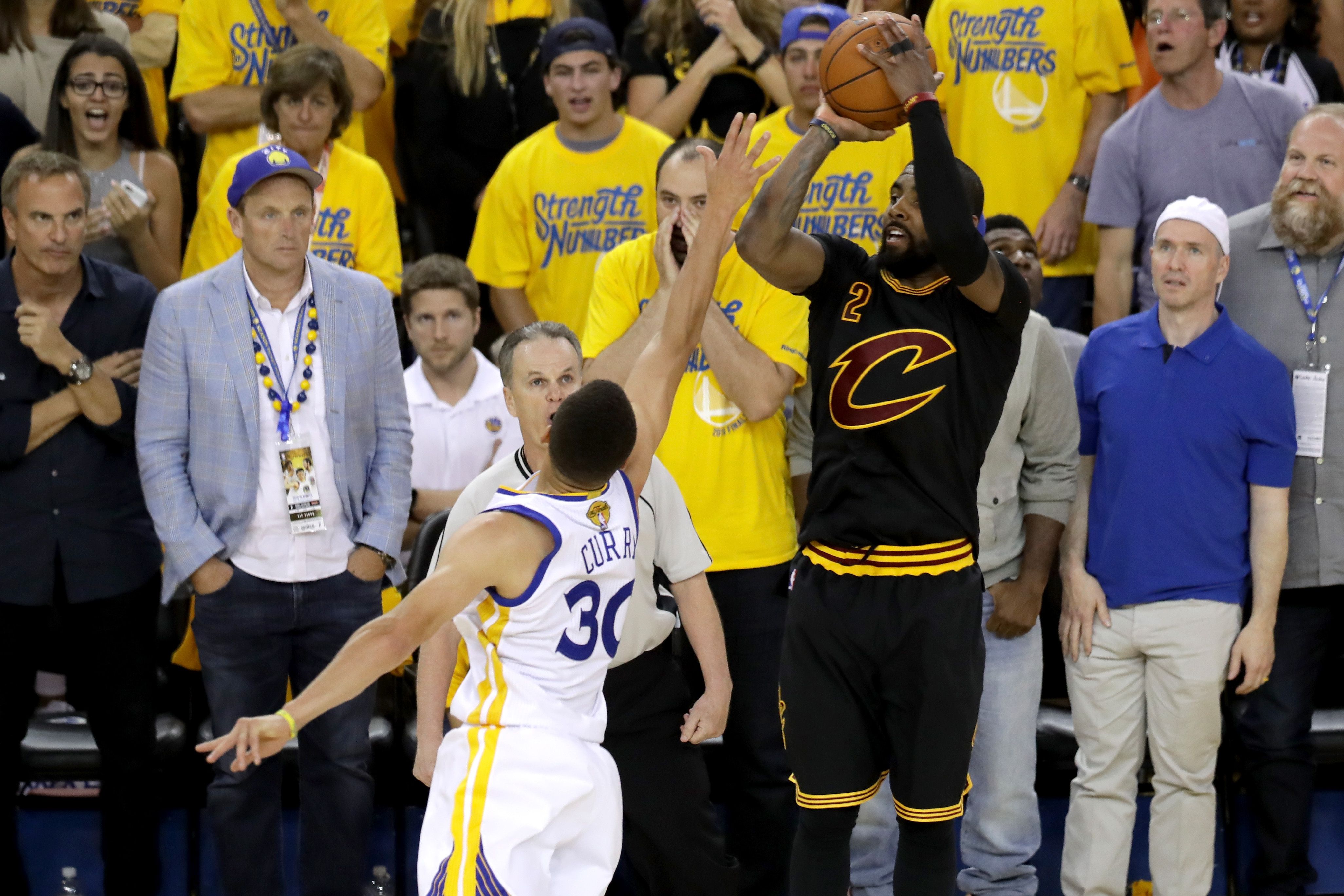 Cavaliers player in black #2 leaps for the ball as Warriors player in white #30 defends; crowd in yellow "Strength in Numbers" shirts cheers in the background.
