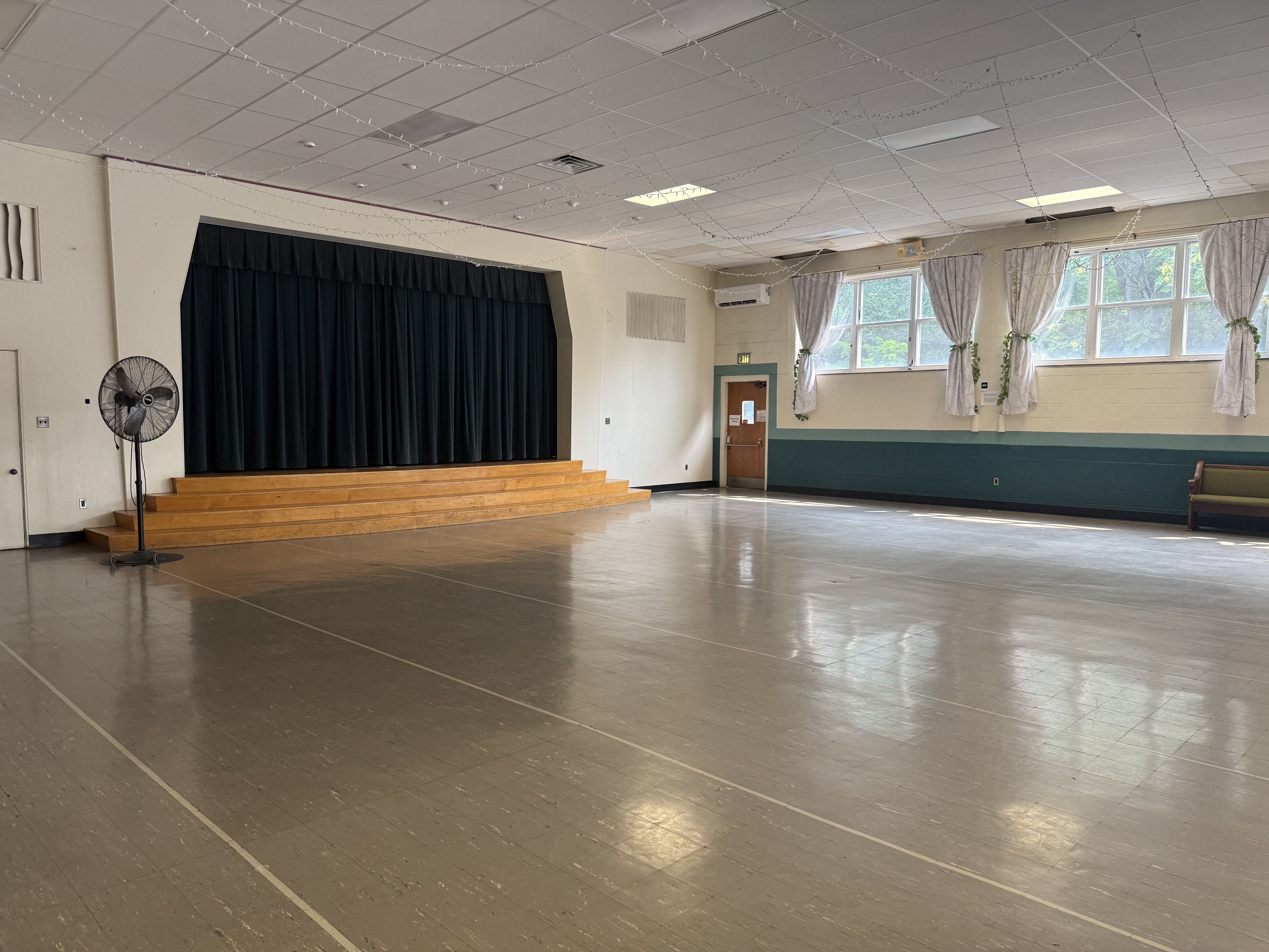 Empty school auditorium with a wooden, stepped stage and dark blue curtains. Ceiling string lights, a standing fan on the left, and large windows with white curtains on the right.