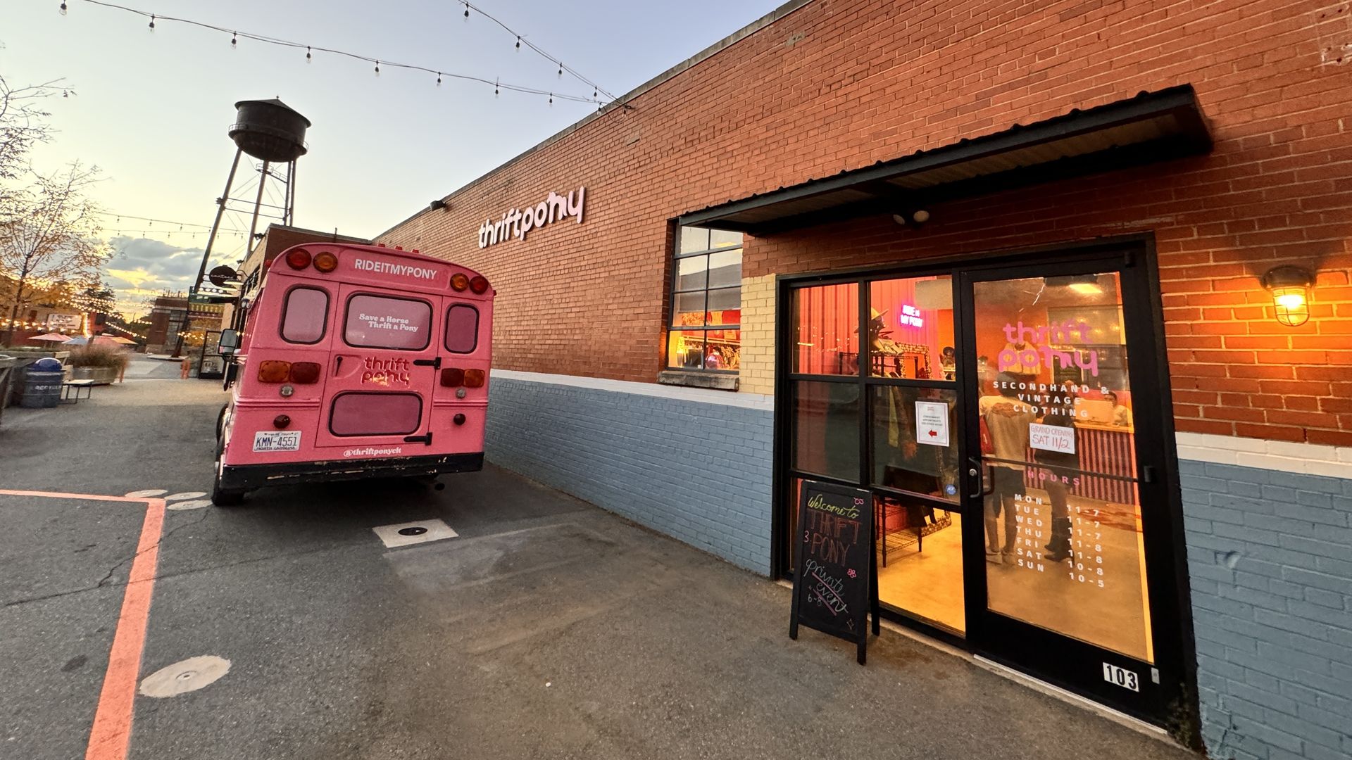 Evening street view of thriftpony's red brick building with a pink thriftpony bus parked outside under string lights, a water tower in the background, and shop hours on the glass door.