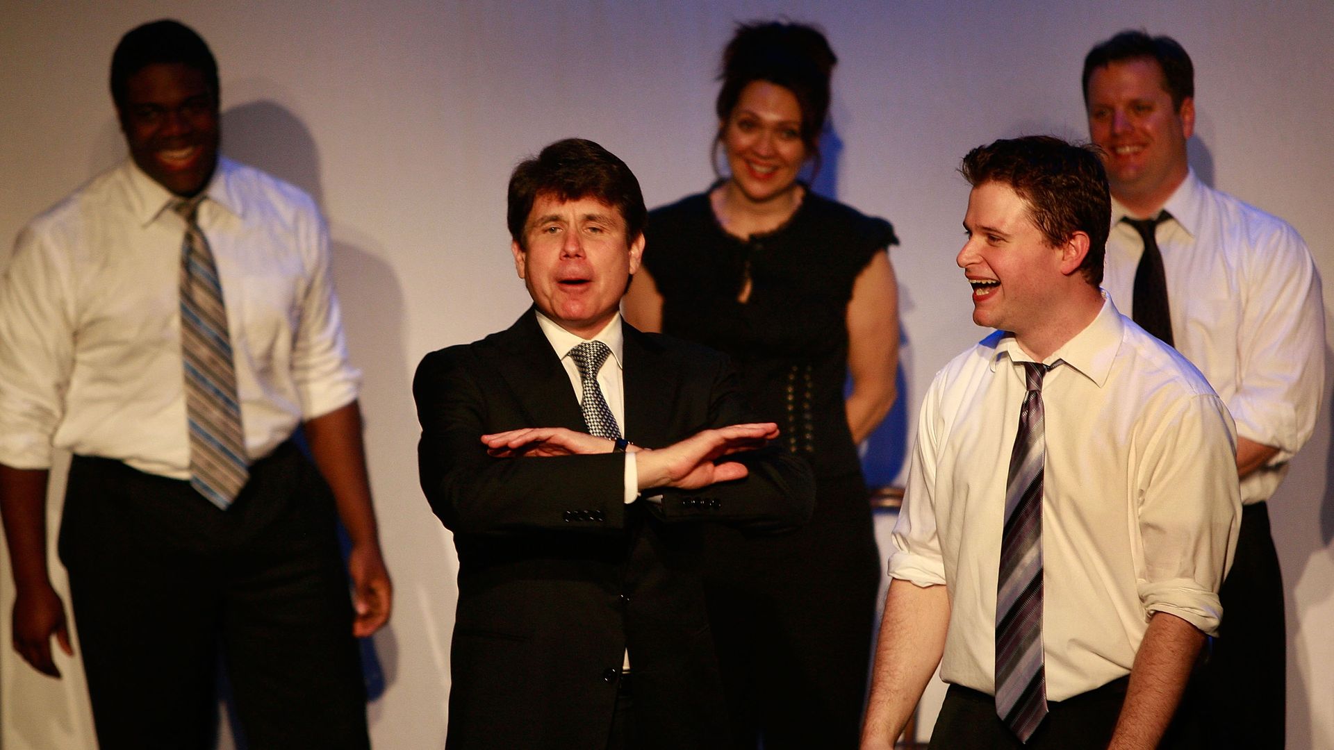 Photo of several people on stage surrounding one man in a suit who is gesturing no while others laugh. 