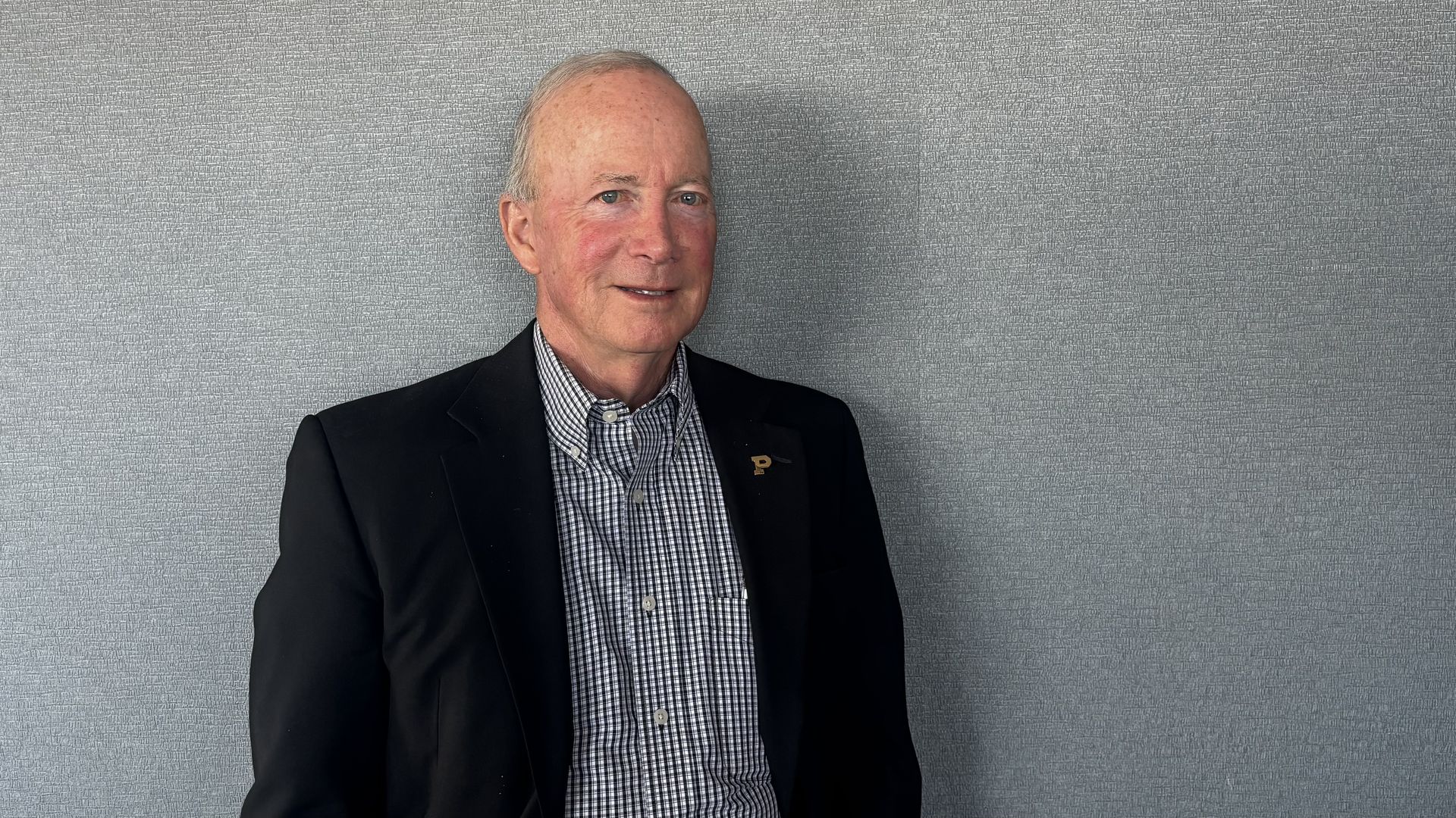 An older man with short gray hair stands against a light gray textured wall, wearing a dark blazer over a checkered shirt and a lapel pin, looking to the left with a mild smile.