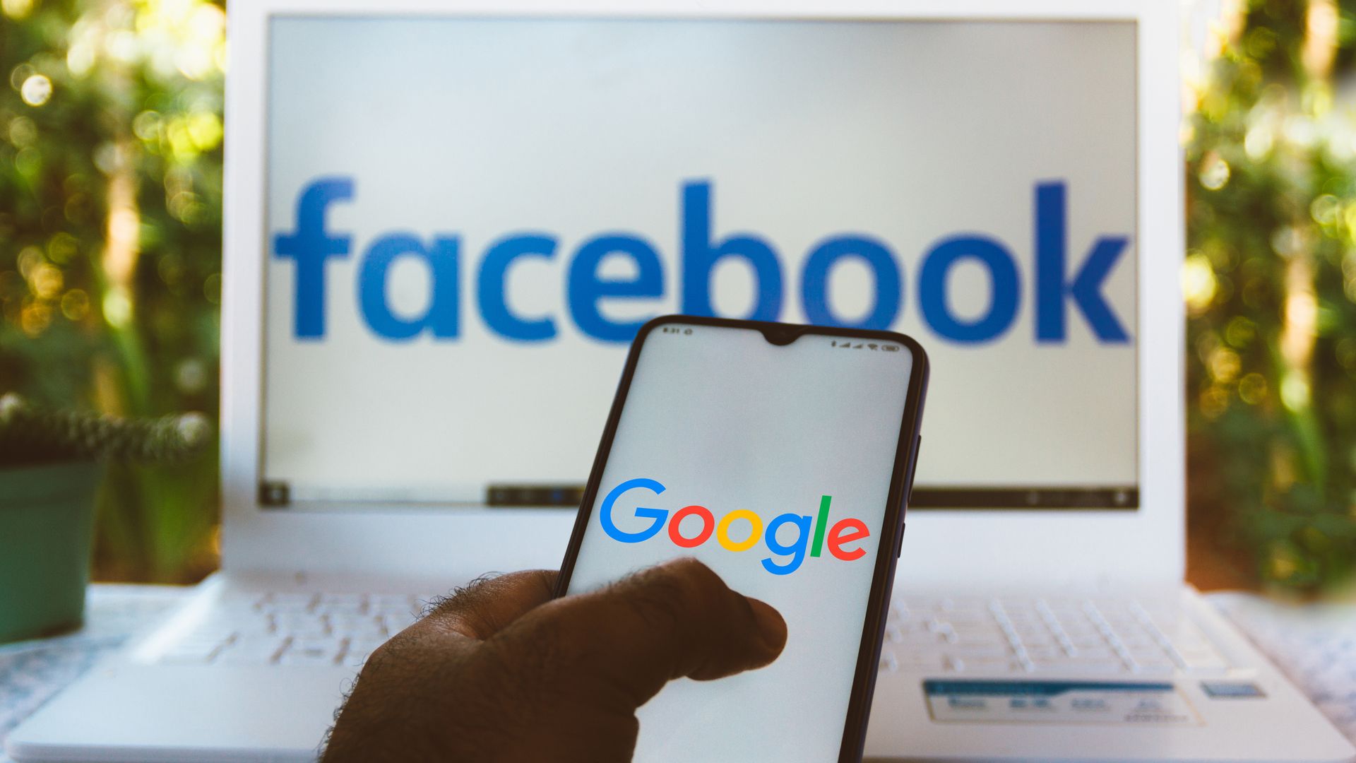 A photo of a smartphone displaying the Google logo, held in front of a laptop displaying the Facebook logo