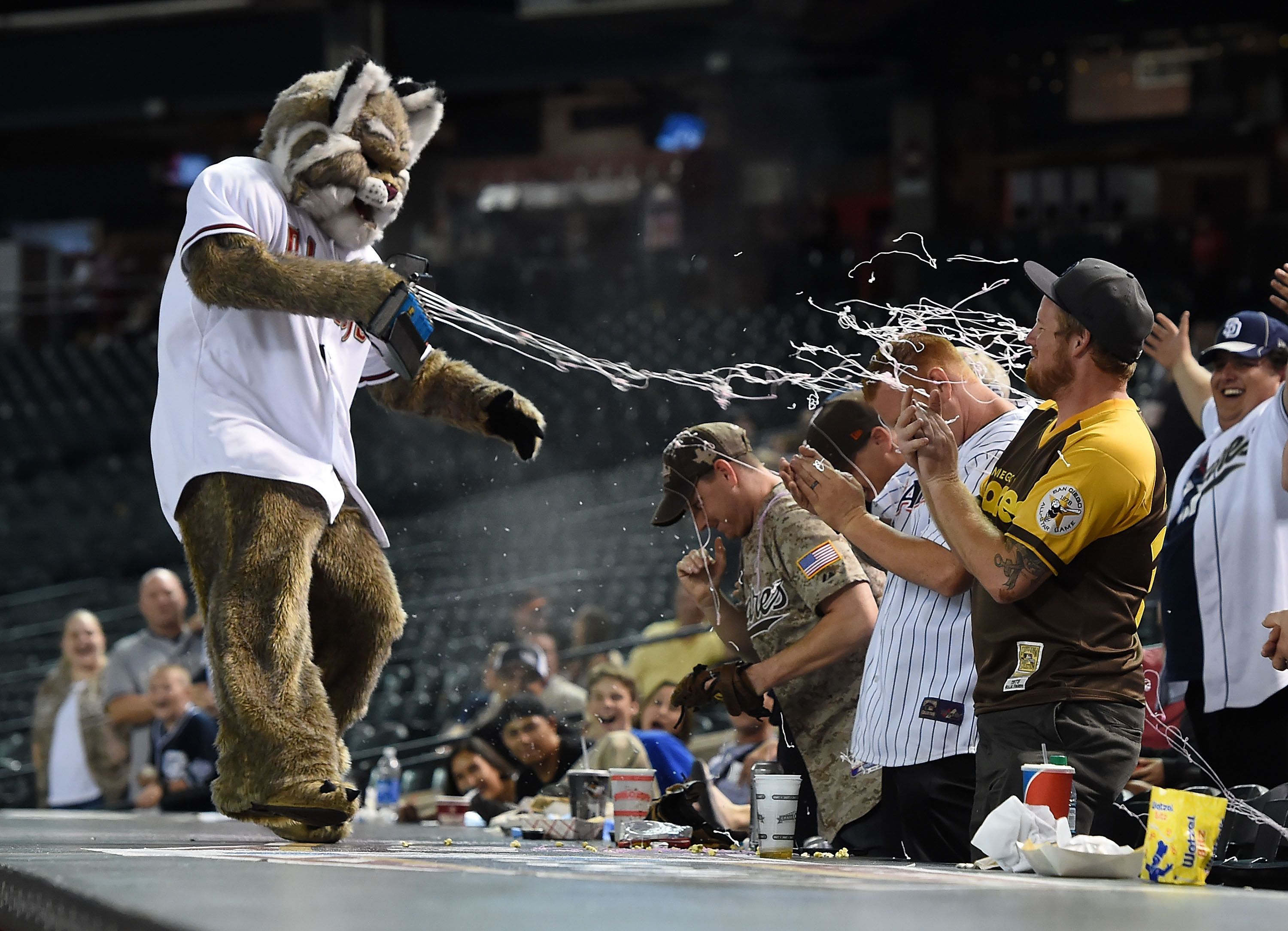 A furry mascot in a white baseball jersey faces cheering fans as stringy confetti shoots across the arena, with players and spectators visible in the background.