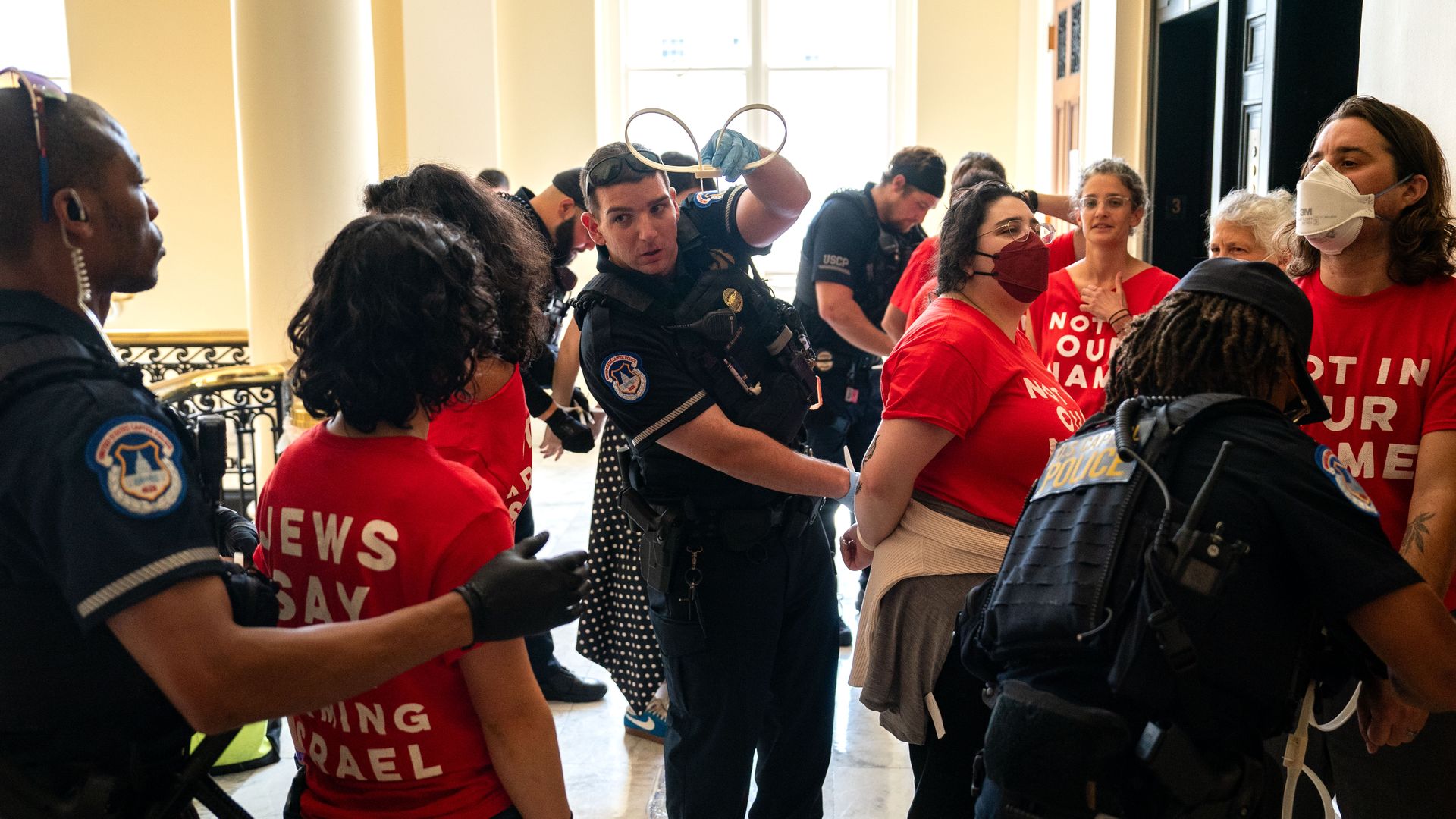 Capitol Police arresting people in red shirts with zipties next to elevators in a white stairwell.