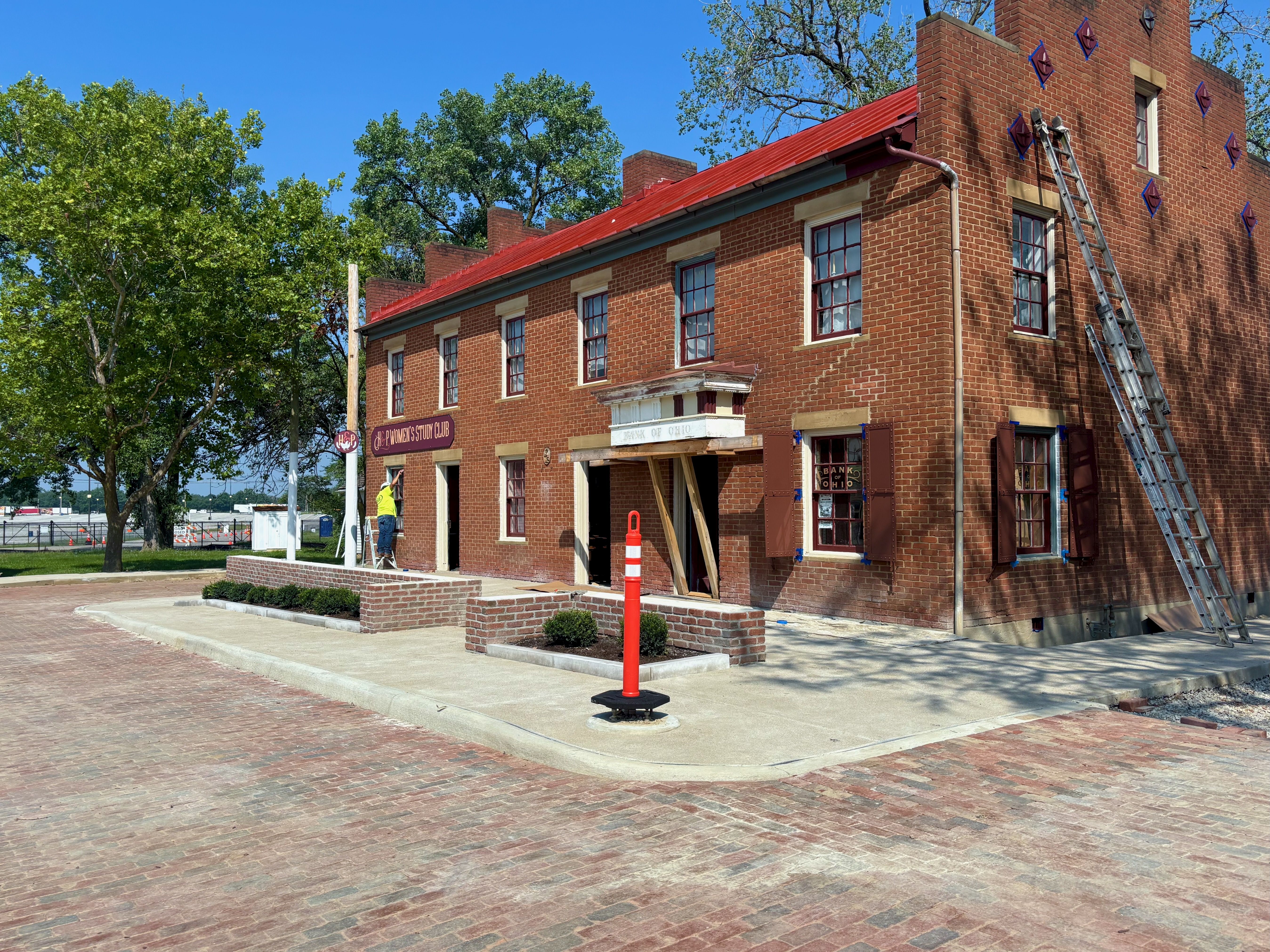 Brick building under renovation with a red metal roof, ladders leaning on the right, a worker in a yellow vest near the entrance, and green trees against a bright blue sky.