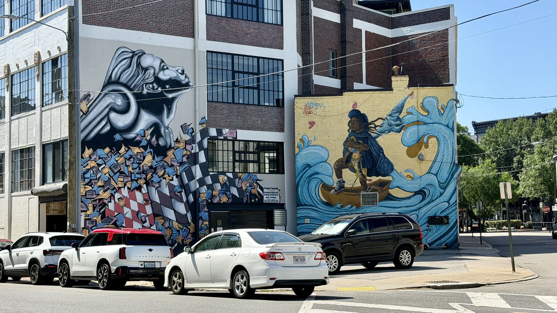 Urban street with two large murals: left shows a grayscale head with floating books; right a yellow brick wall depicting a girl amid blue waves. Cars line the curb under a clear blue sky.