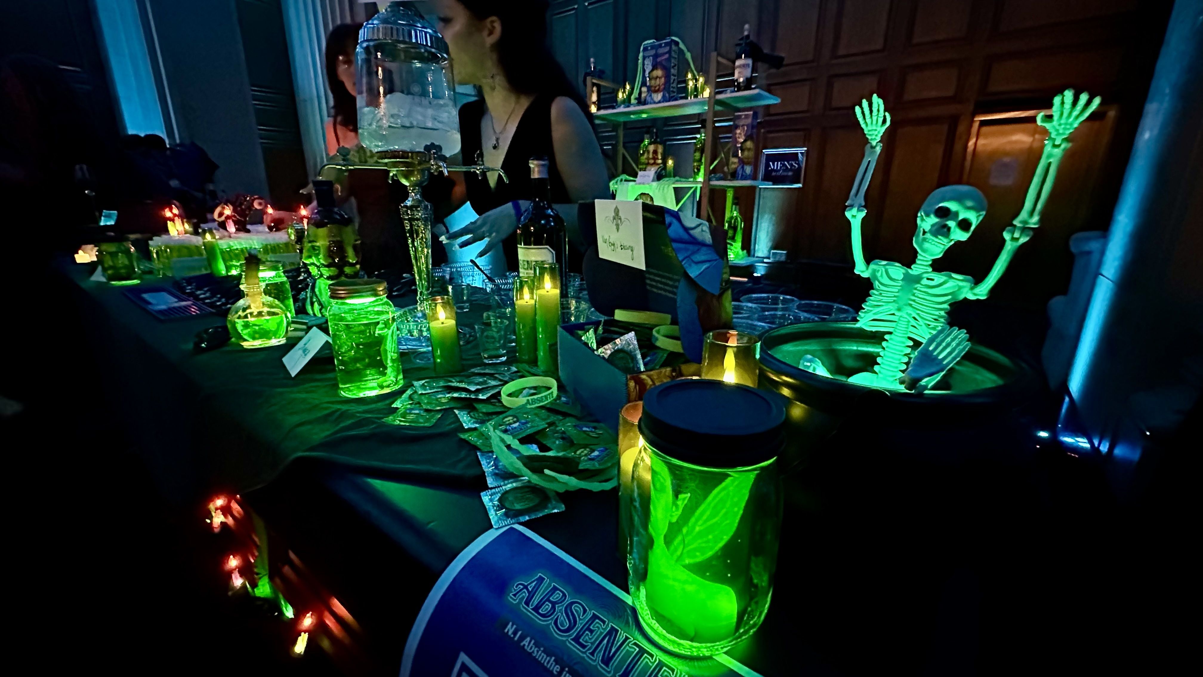 Glow-in-the-dark green absinthe bottles and candles on black tablecloth at a dimly lit event, with a small glowing skeleton figure raising its arms from a cauldron and people in the background.