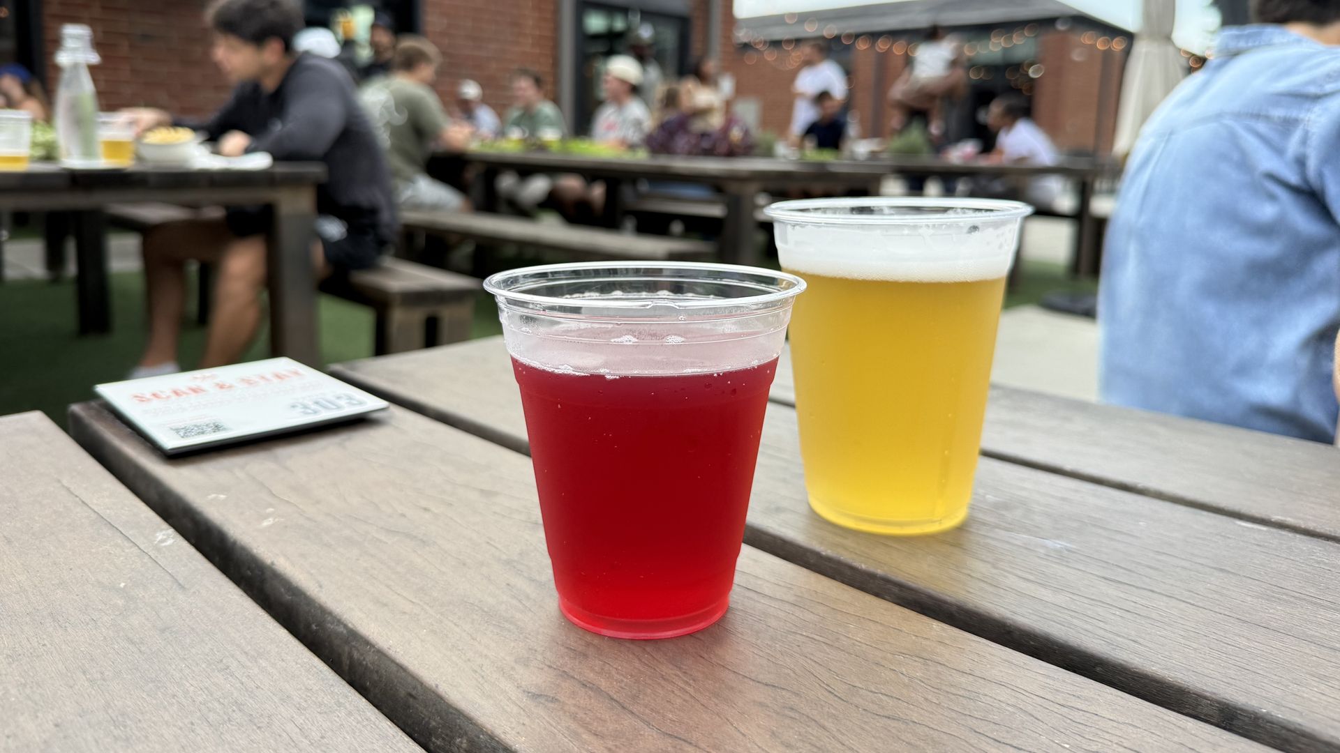 Outdoor beer garden with two plastic cups in the foreground: red beverage and yellow beer, on a wooden table; a blurred group sits at picnic tables under string lights by brick buildings.