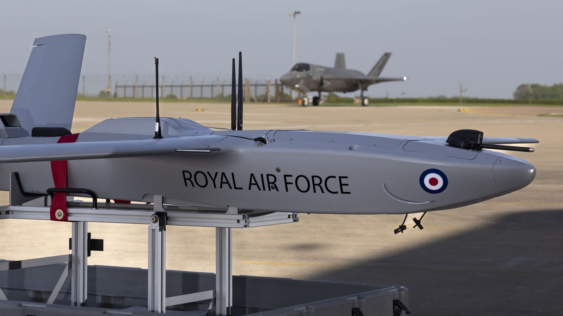 A grey-colored drone sits in the foreground. An F-35 aircraft sits in the background. They are both on the tarmac.