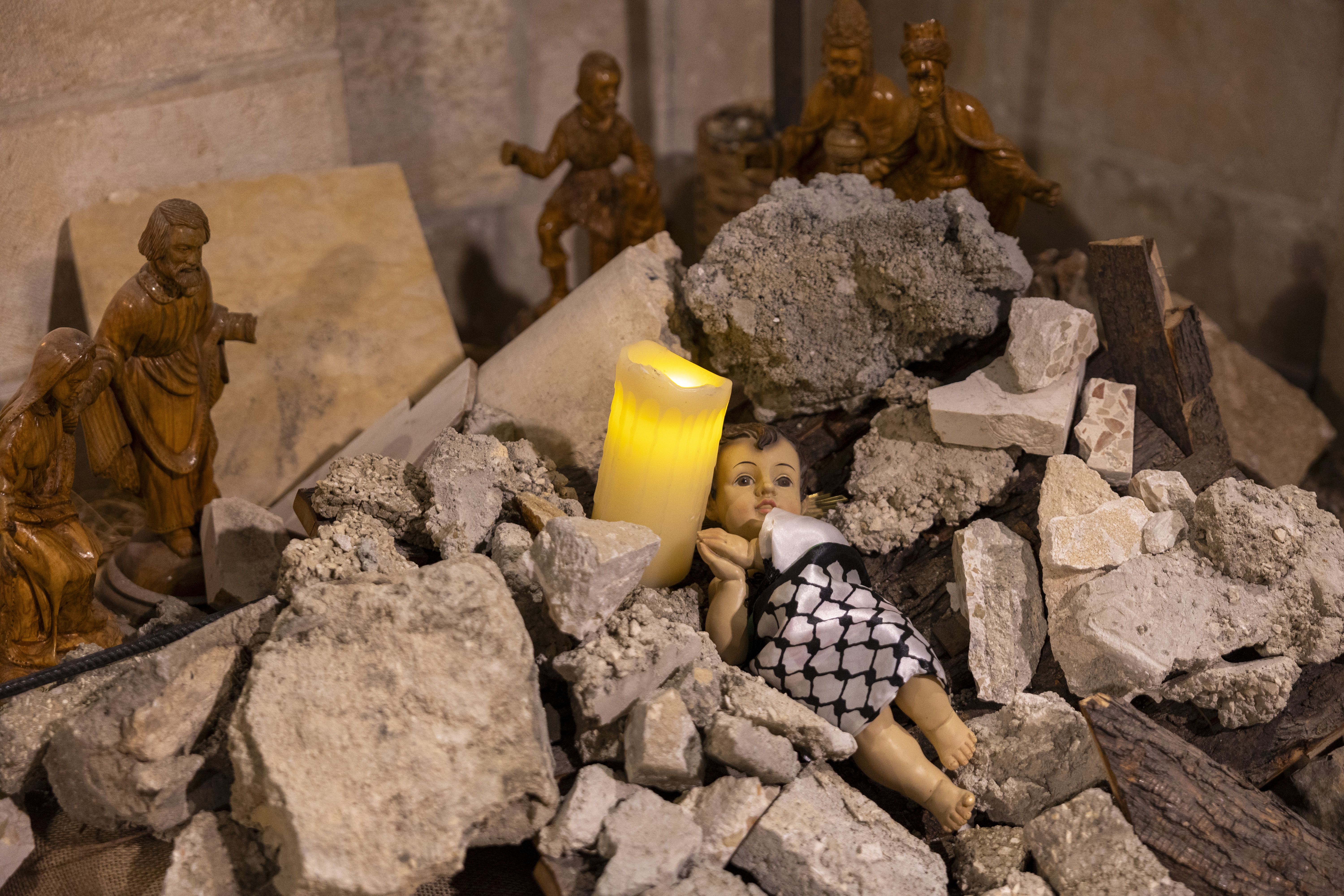 A plastic baby doll wrapped in a keffiyah scarf placed among rubble next to an electric candle. 