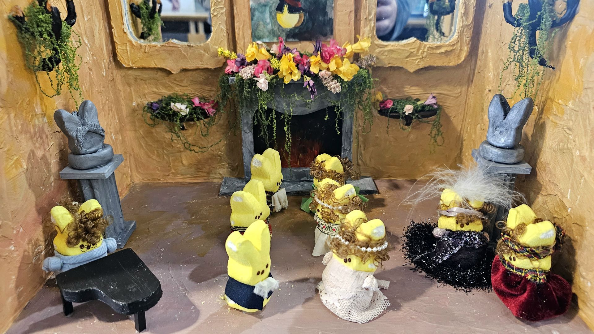 Whimsical diorama with yellow plush bunnies in dresses gathered around a lit fireplace under a flower-filled mantle; gray bunny statues on pedestals flank orange, textured walls.