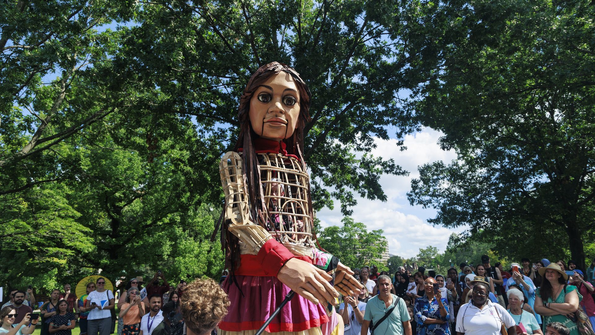 An oversized puppet of a girl towers over a crowd of people gathered in an area shaded by trees.