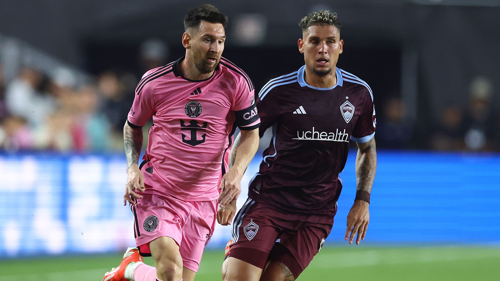 Two soccer players run forward, with one man wearing a pink jersey and the other wearing a burgundy-colored jersey.
