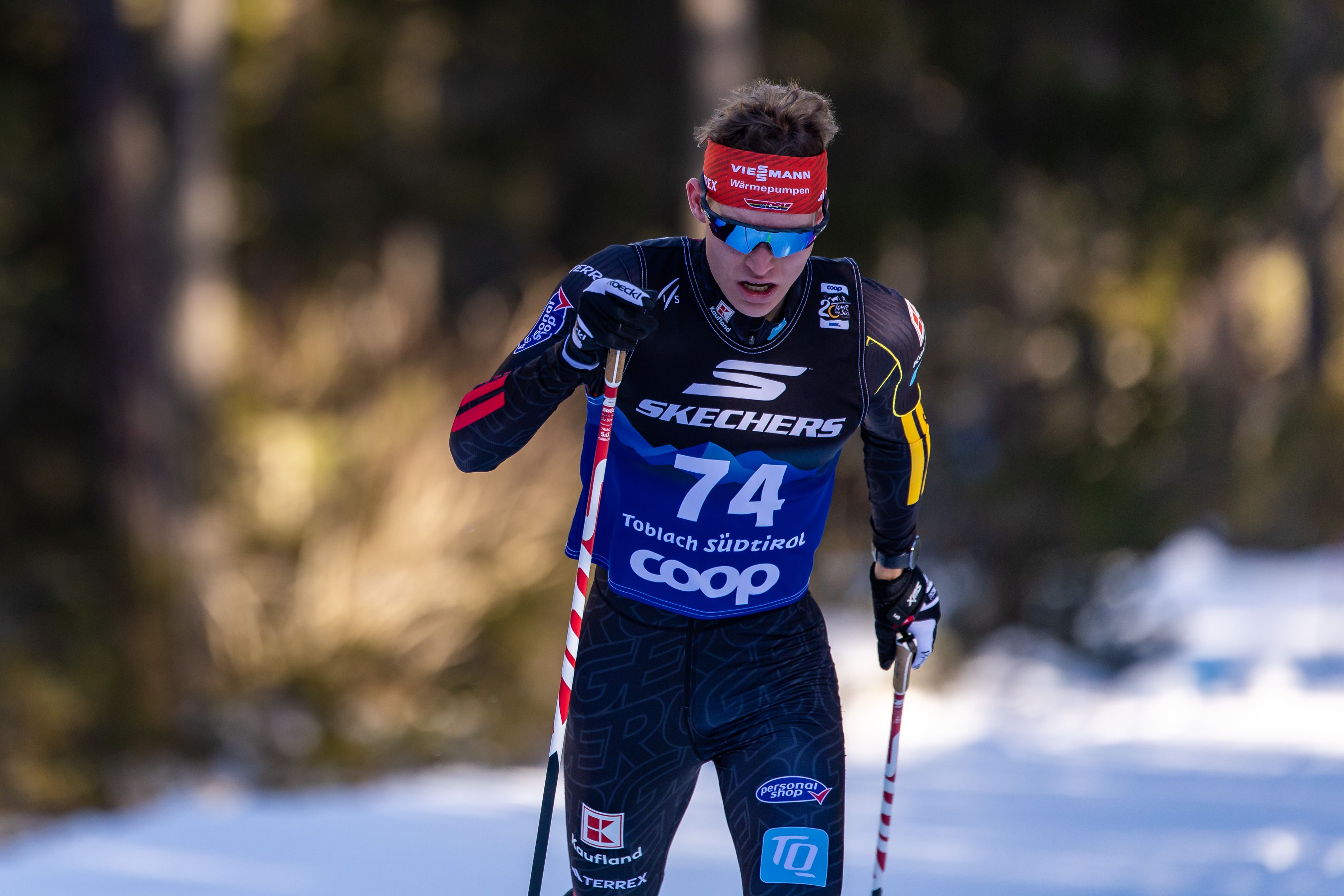 Cross-country skier wearing black and red gear with bib number 74, skiing on a snowy trail surrounded by trees, wearing blue reflective sunglasses and a red headband.