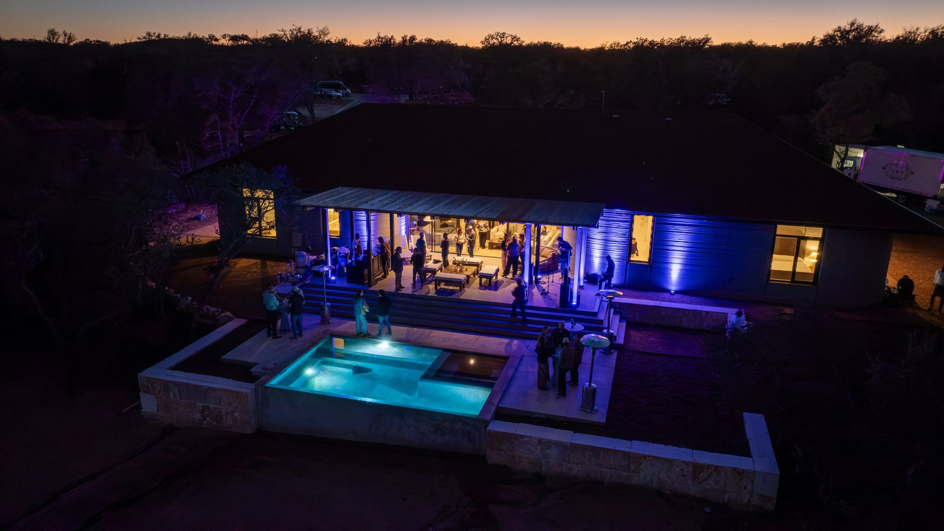 Evening outdoor gathering at a modern house with blue-lit exterior walls and a glowing blue swimming pool, people socializing on the patio and around the pool under sunset sky.
