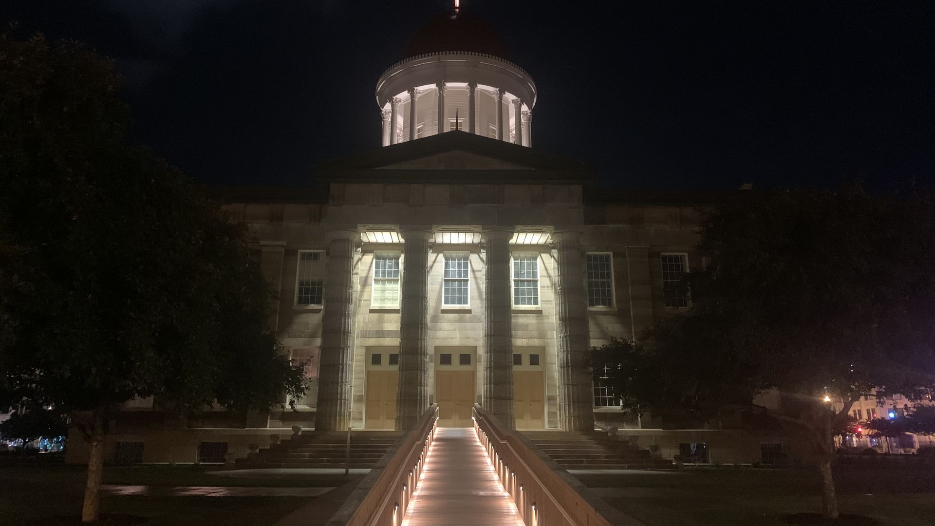 Night view of a domed, neoclassical building with tall columns. A bright walkway leads to the entrance, flanked by trees. An American flag flies atop the dome against a dark sky.