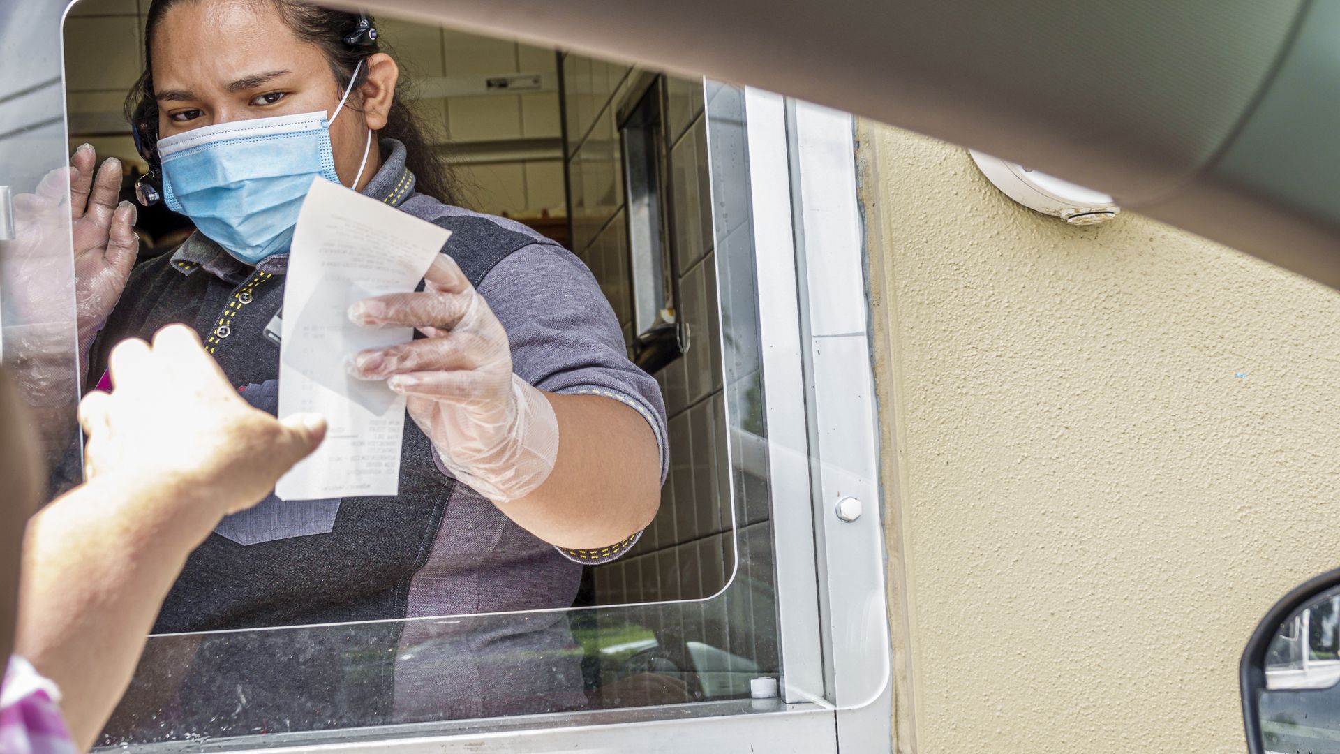 Photo of a masked worker with one arm reaching out a window with a receipt toward a hand in the car