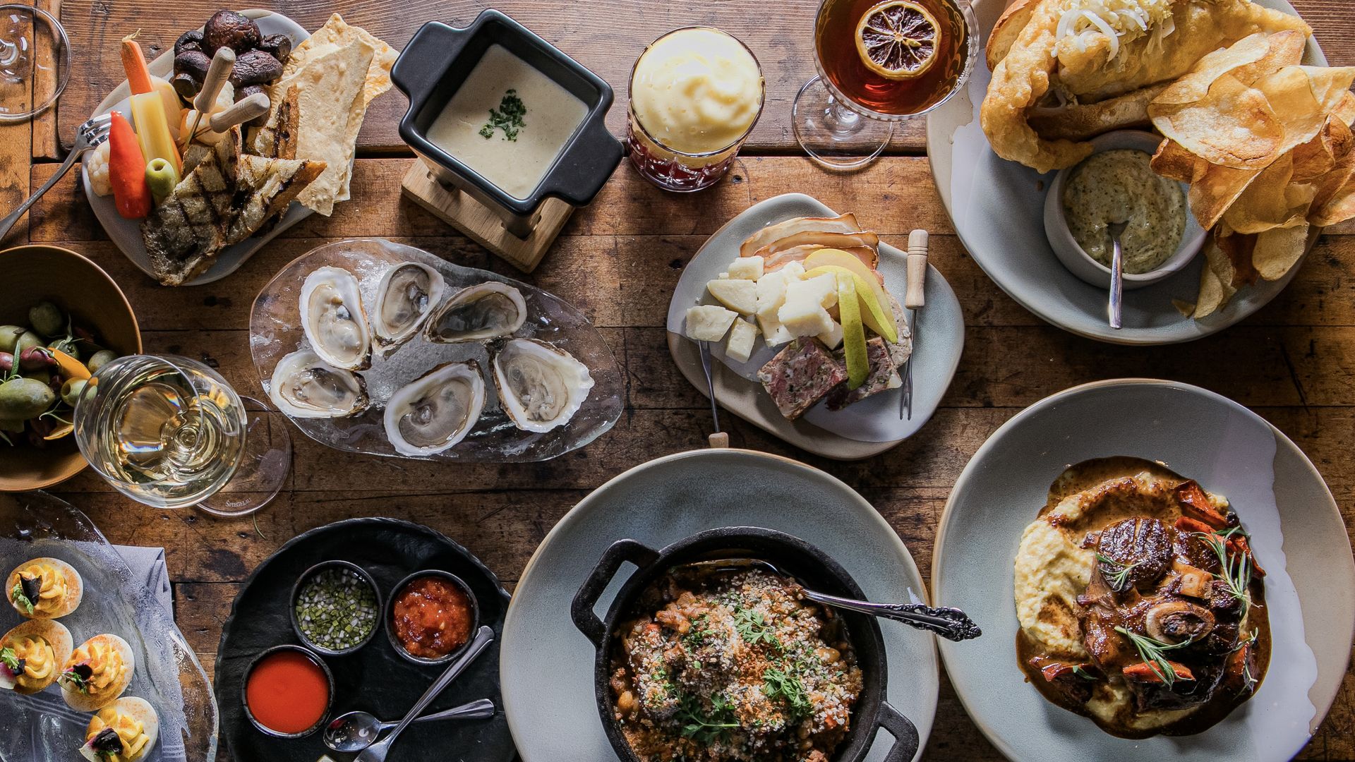 Overhead view of a rustic wooden table with diverse dishes including oysters, cheese and meat platter, deviled eggs, olives, hot sauces, a creamy stew, fish and chips with chips, and drinks with lemon and foam.