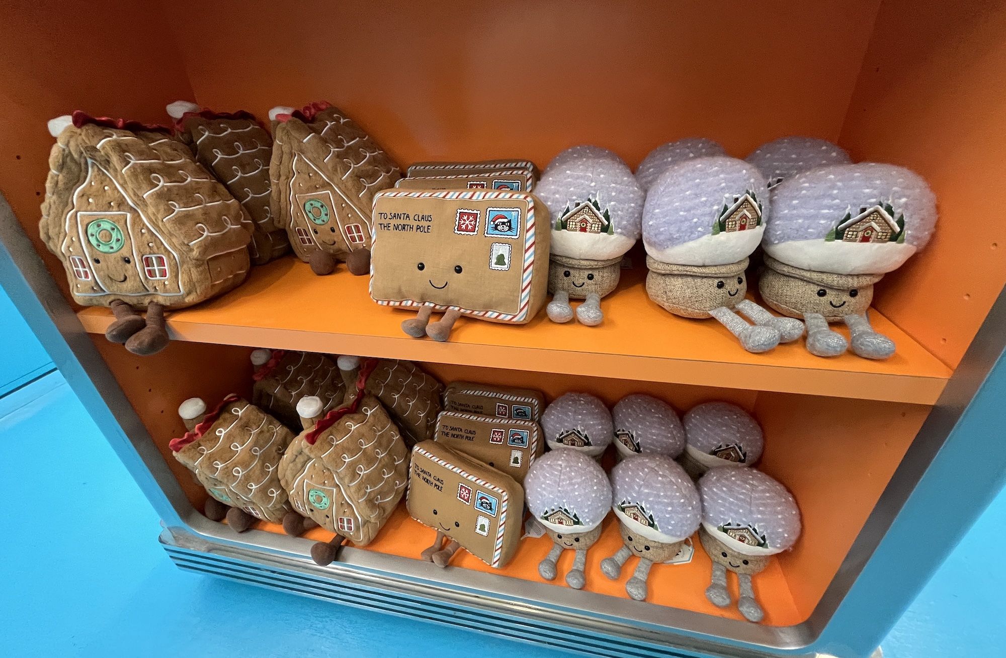 Jellycat plush holiday toys on orange shelves: gingerbread houses, letters to Santa with faces, and snow globes with smiling faces and cozy cottages inside.