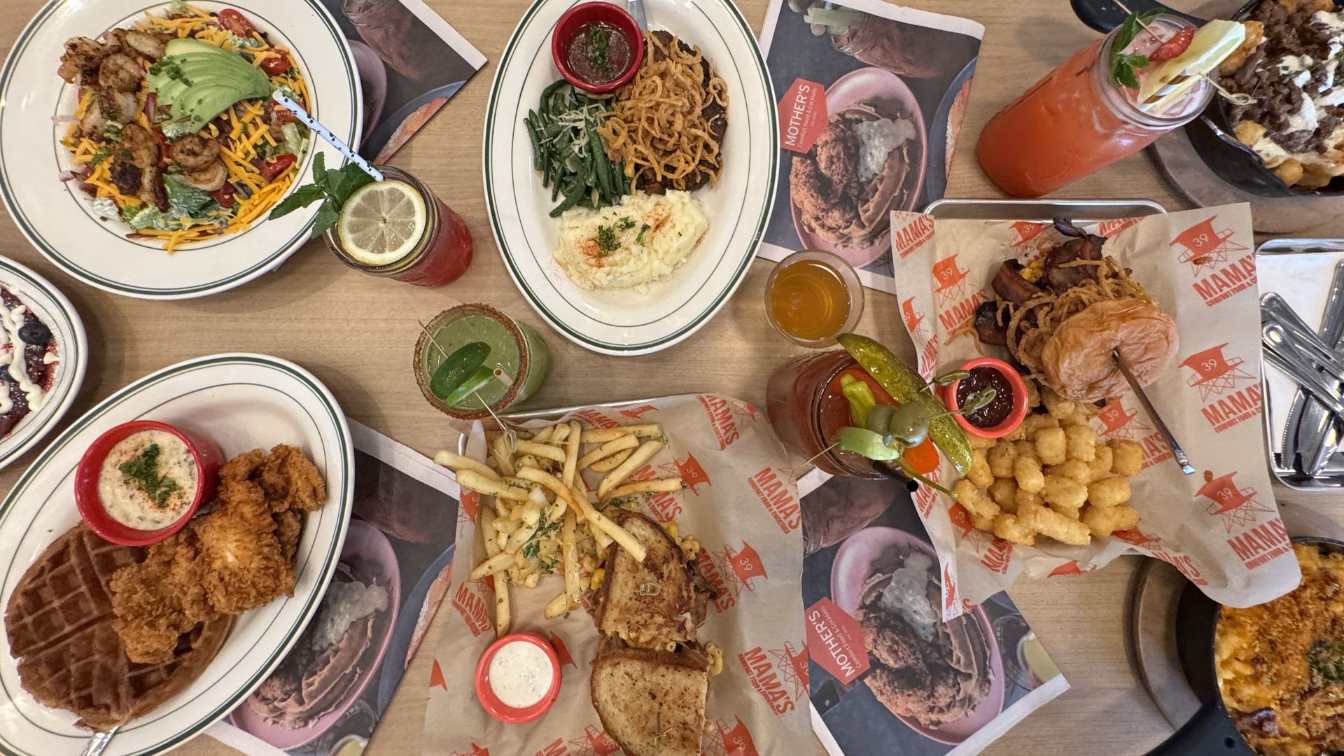 A table with dishes including shrimp salad with avocado, mashed potatoes and green beans, fried chicken and waffles, sandwich with fries, and two cocktails, one garnished with lemon, one with pickles.