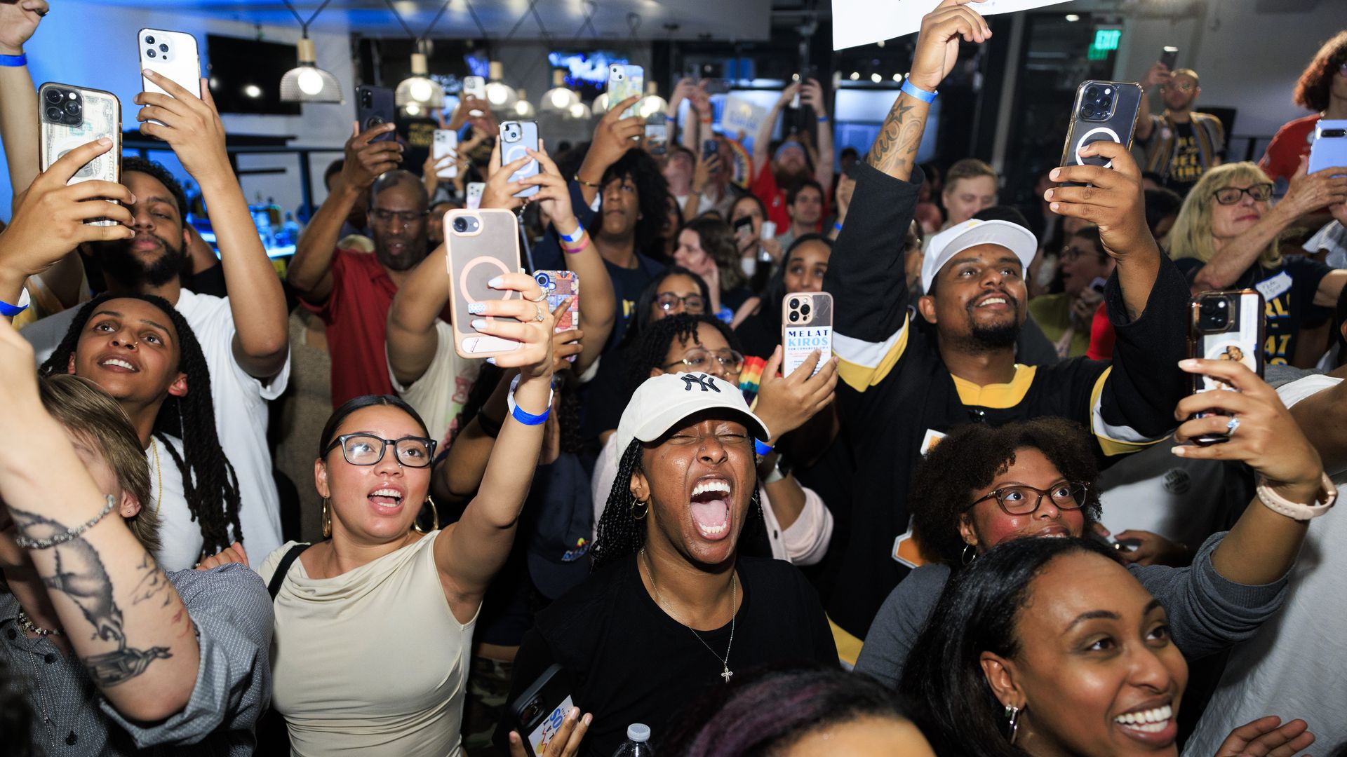 Large crowd at an indoor event, everyone holding up phones to take selfies and photos; blue ambient lighting, diverse group wearing casual clothes and wristbands, excited expressions.