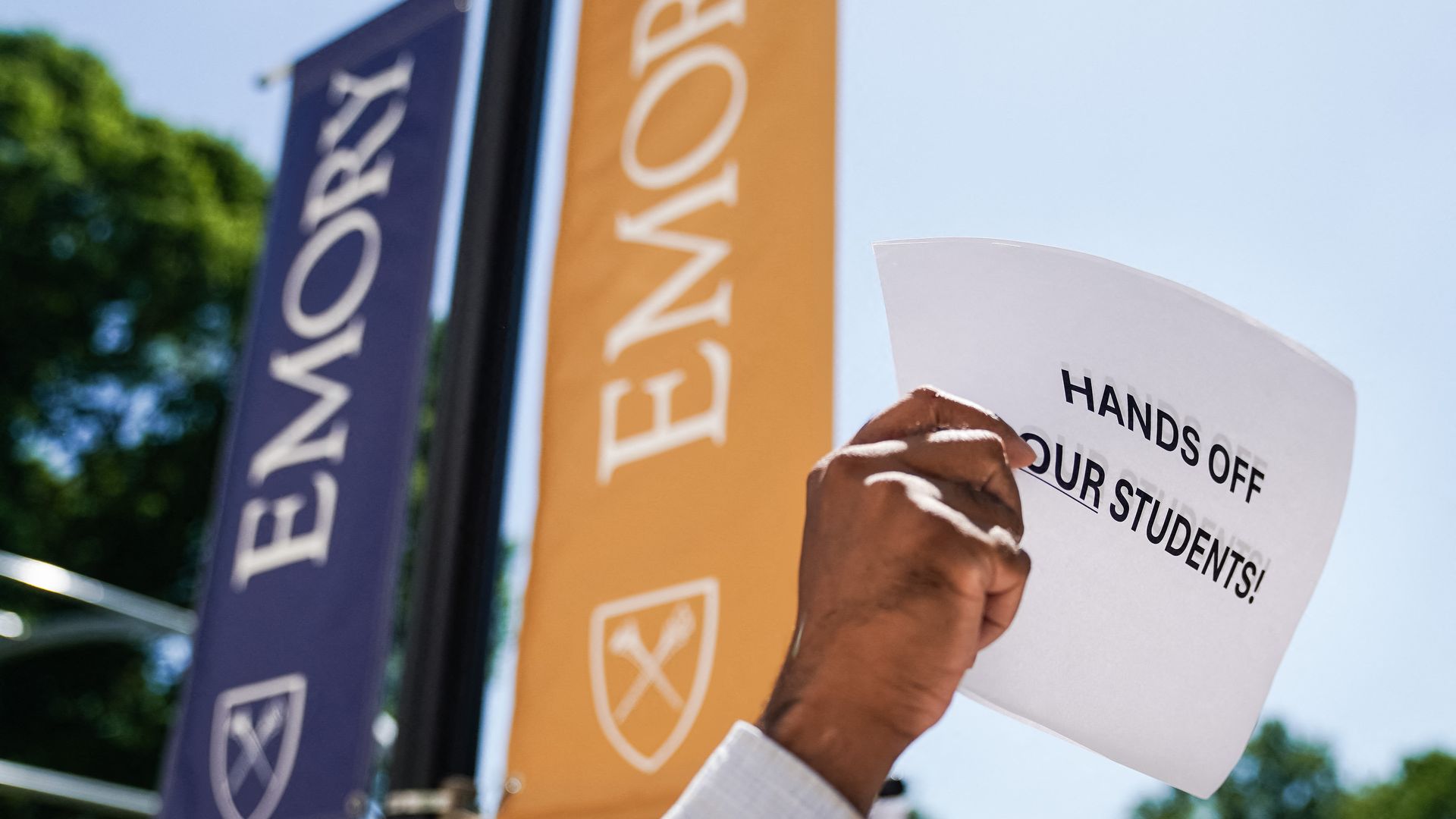 A photo of a hand holding a "Hands off our students!" sign in front of Emory University lamppost banners