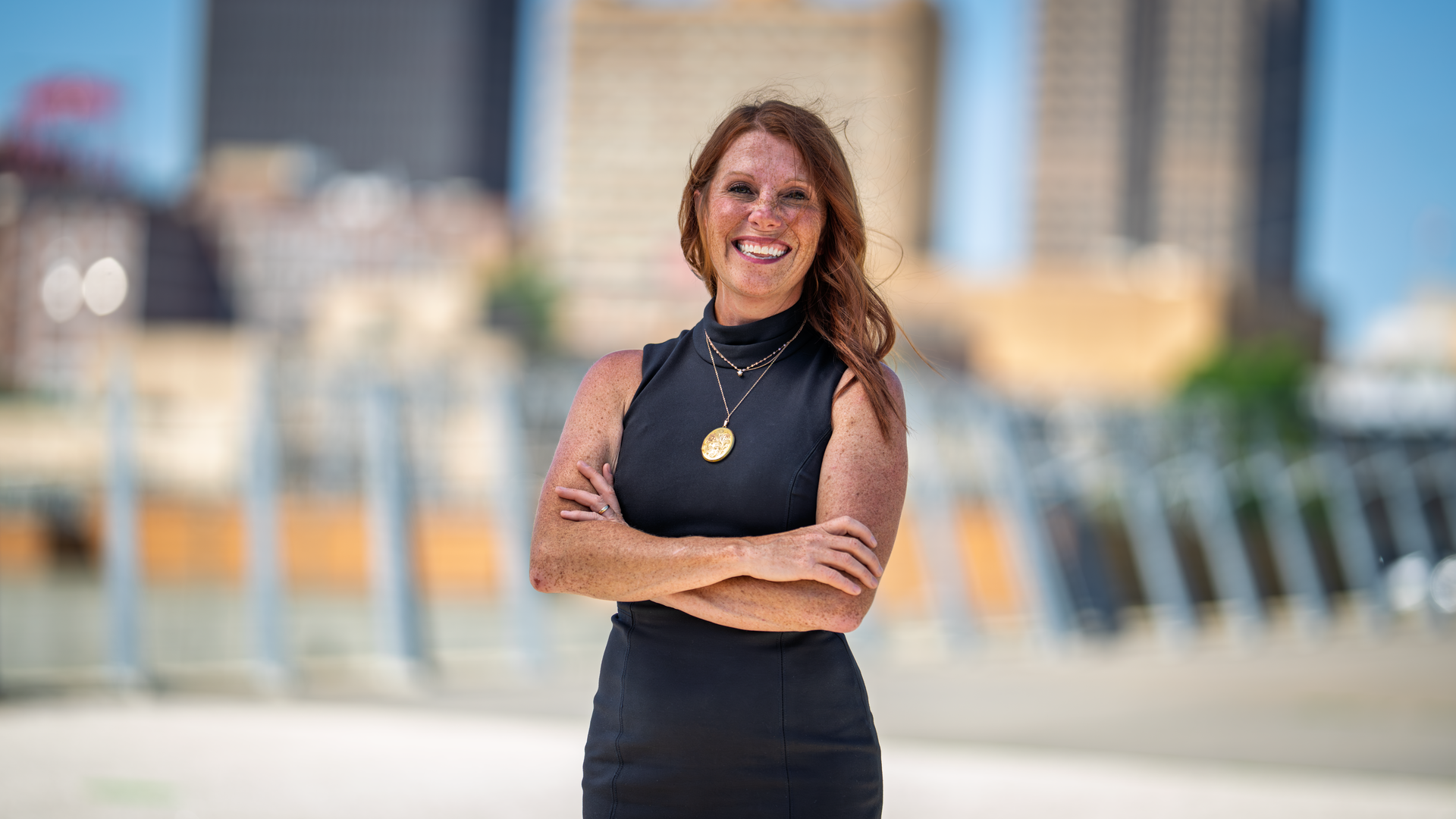 Smiling red-haired woman in a black sleeveless dress stands outdoors with arms crossed, a blurred city skyline behind her. She wears layered necklaces and a gold pendant.