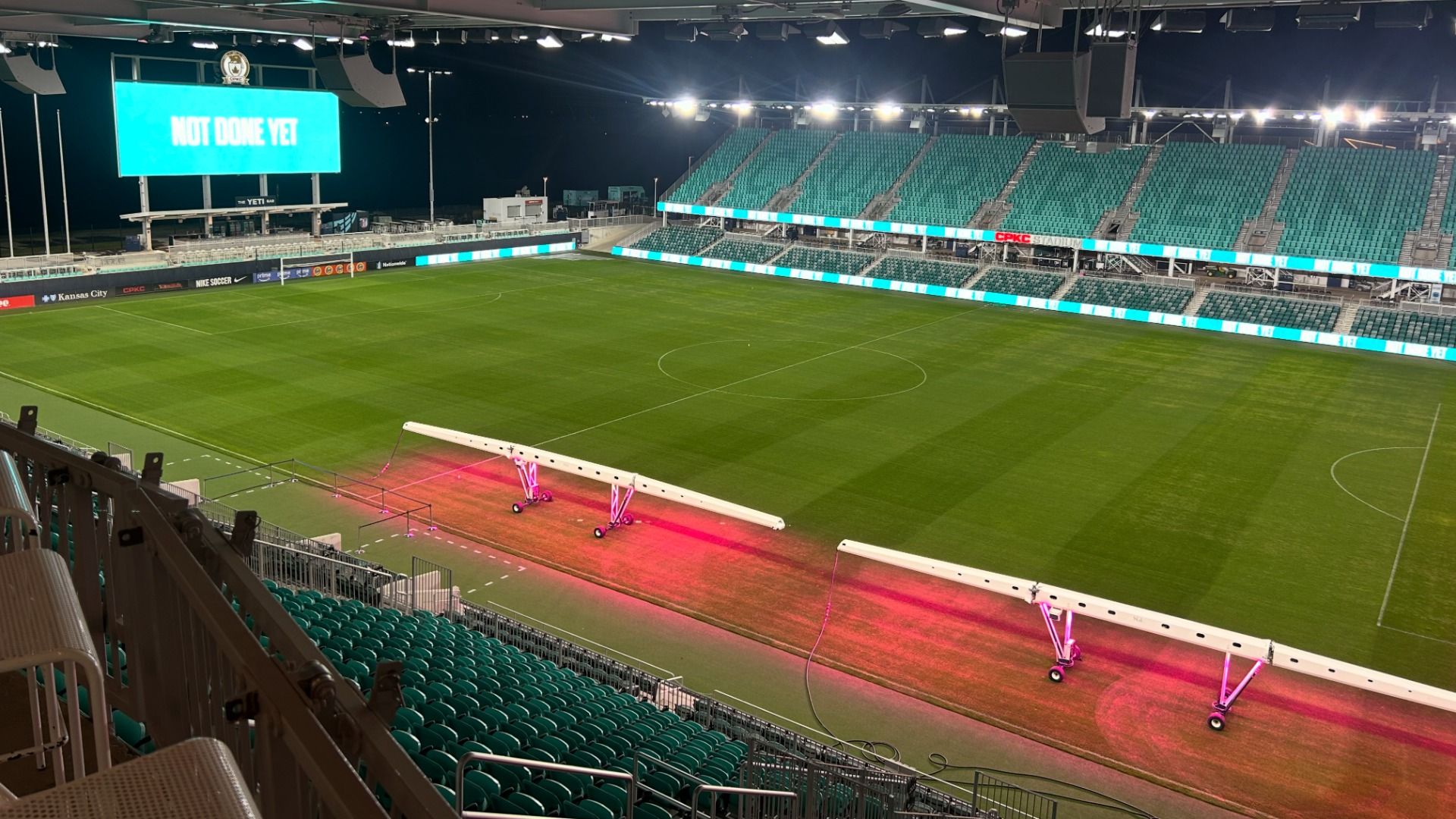 Night view of an empty soccer stadium with bright turquoise seats, a large LED screen saying "NOT DONE YET", and pink-lit maintenance equipment along the sideline on a green pitch.