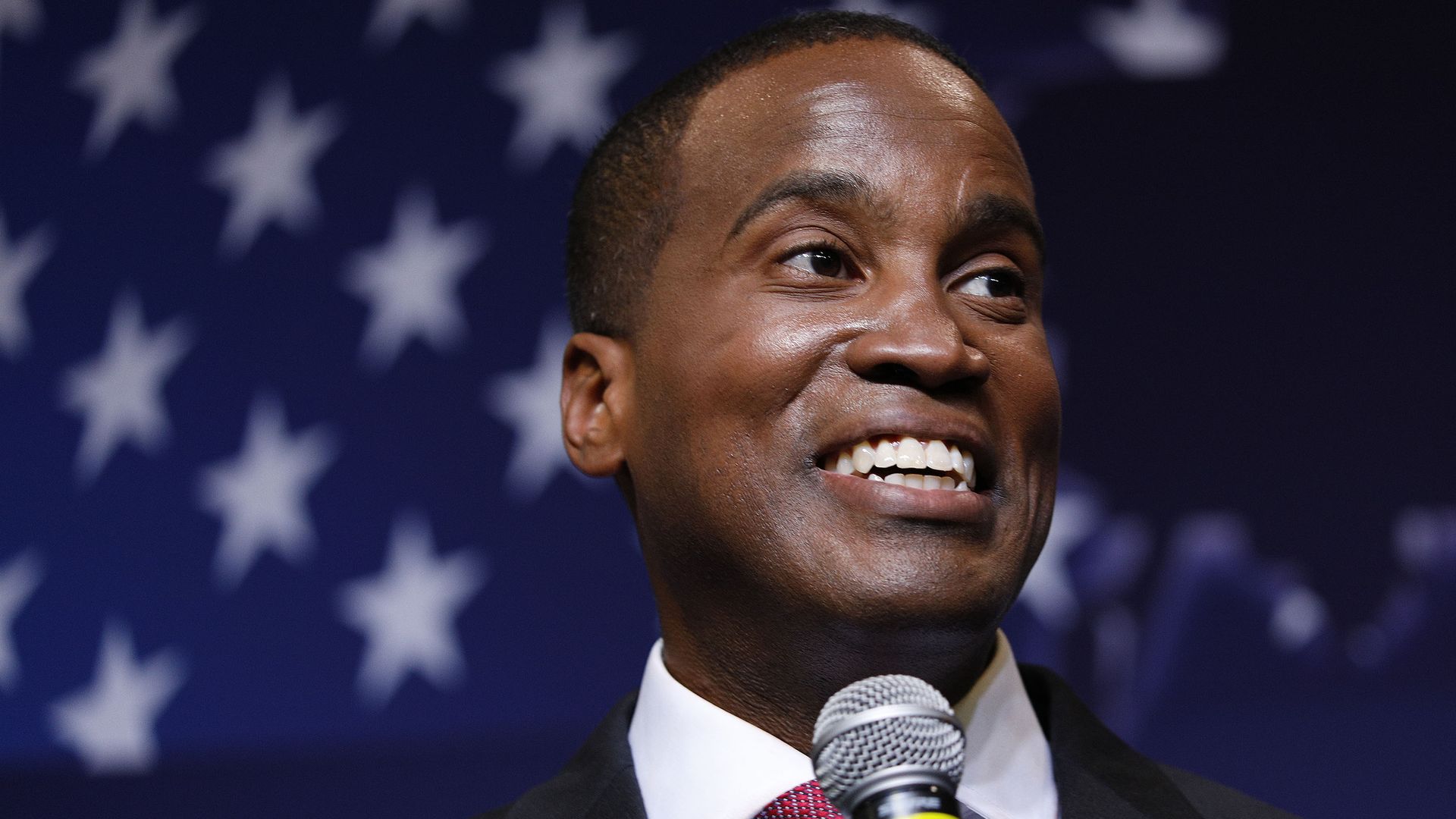 John James holding a microphone with an American flag in the background.