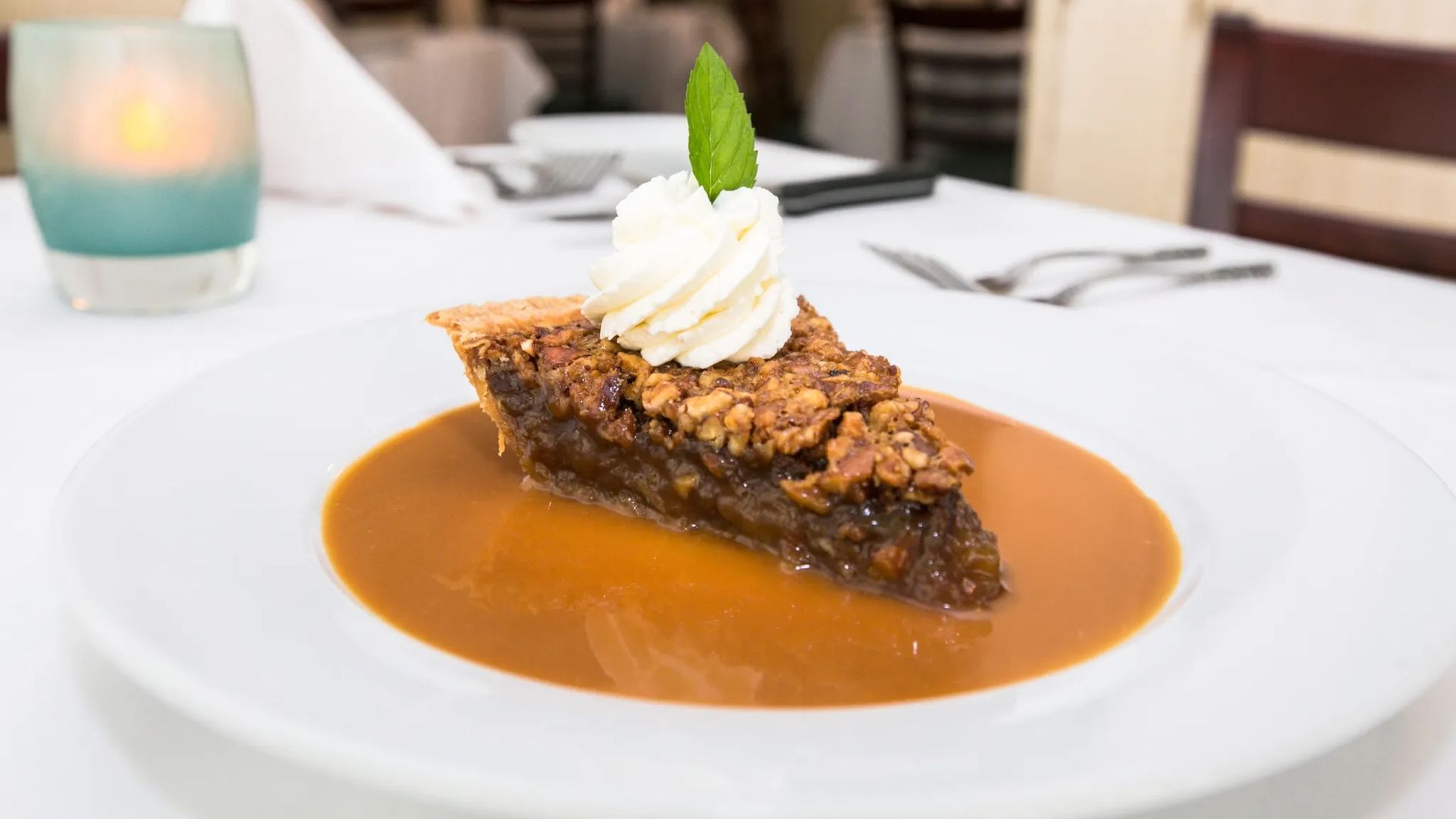Slice of pecan pie topped with whipped cream and a mint leaf, served on a white plate with caramel sauce, set on a table in a softly lit restaurant.