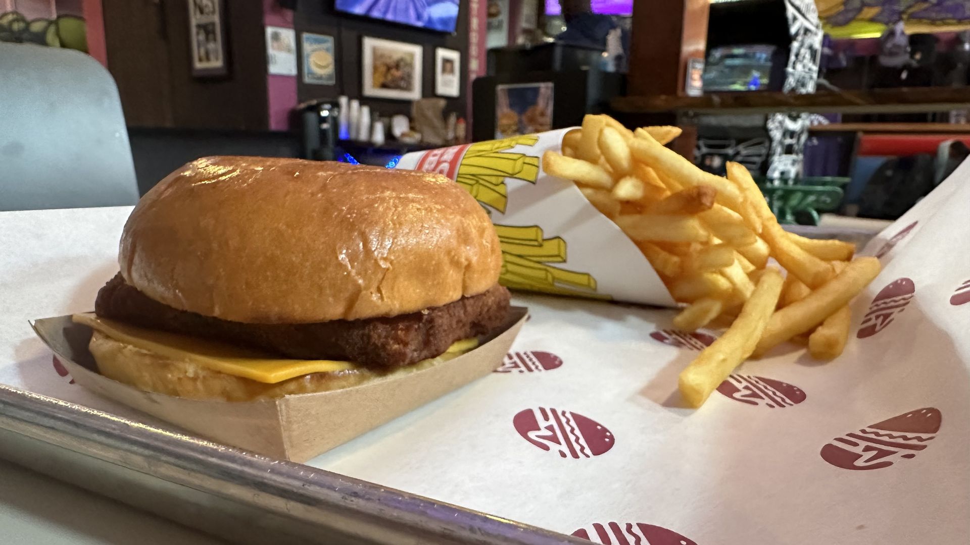 A metal tray is covered with a sheet of wax paper. A fried fish sandwich and a cup of fries are on top of the paper. Arcade games are seen in the background.