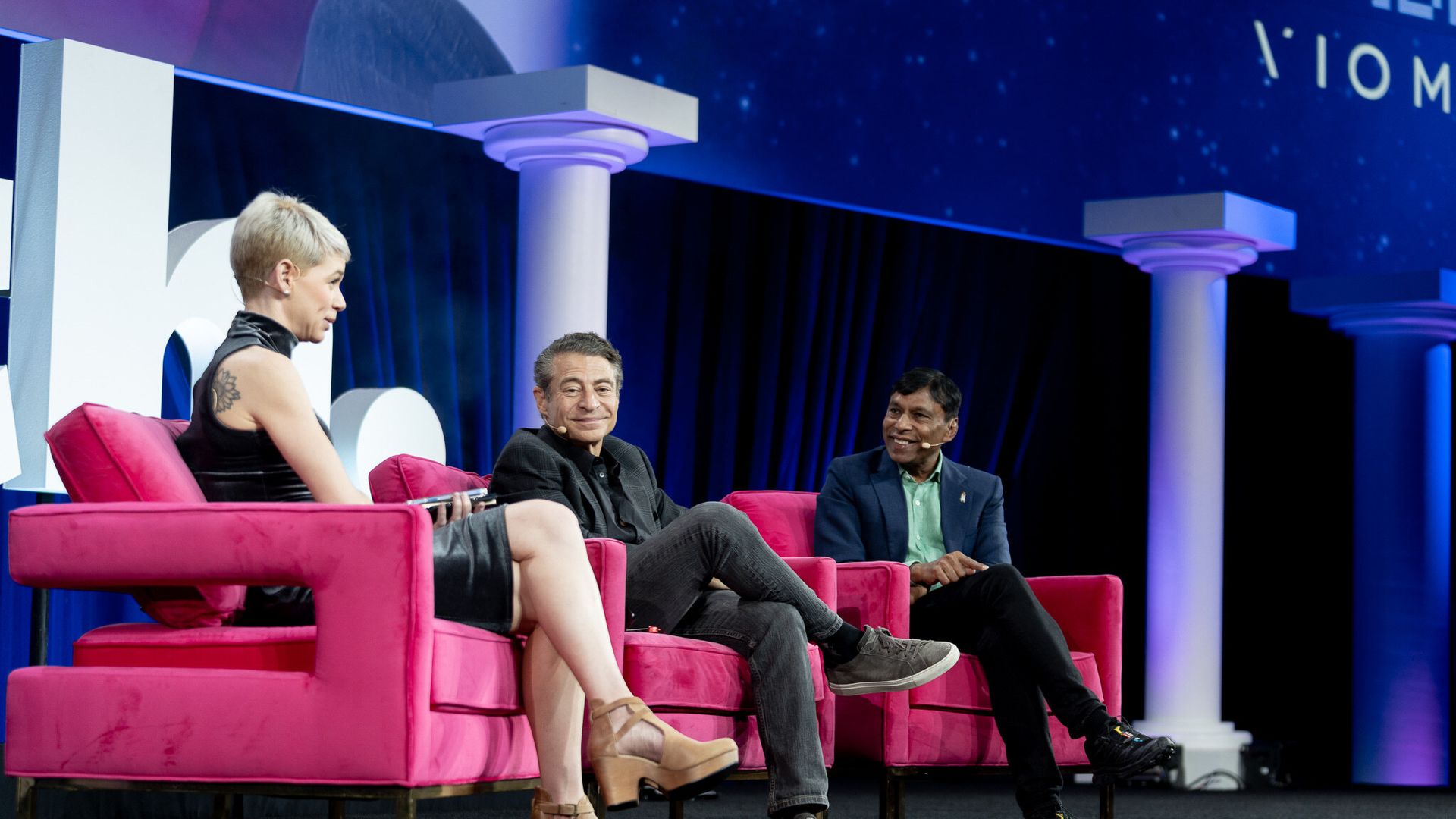 Three people seated on bright pink chairs on stage during a panel discussion, with blue and purple lighting and white columns in the background.
