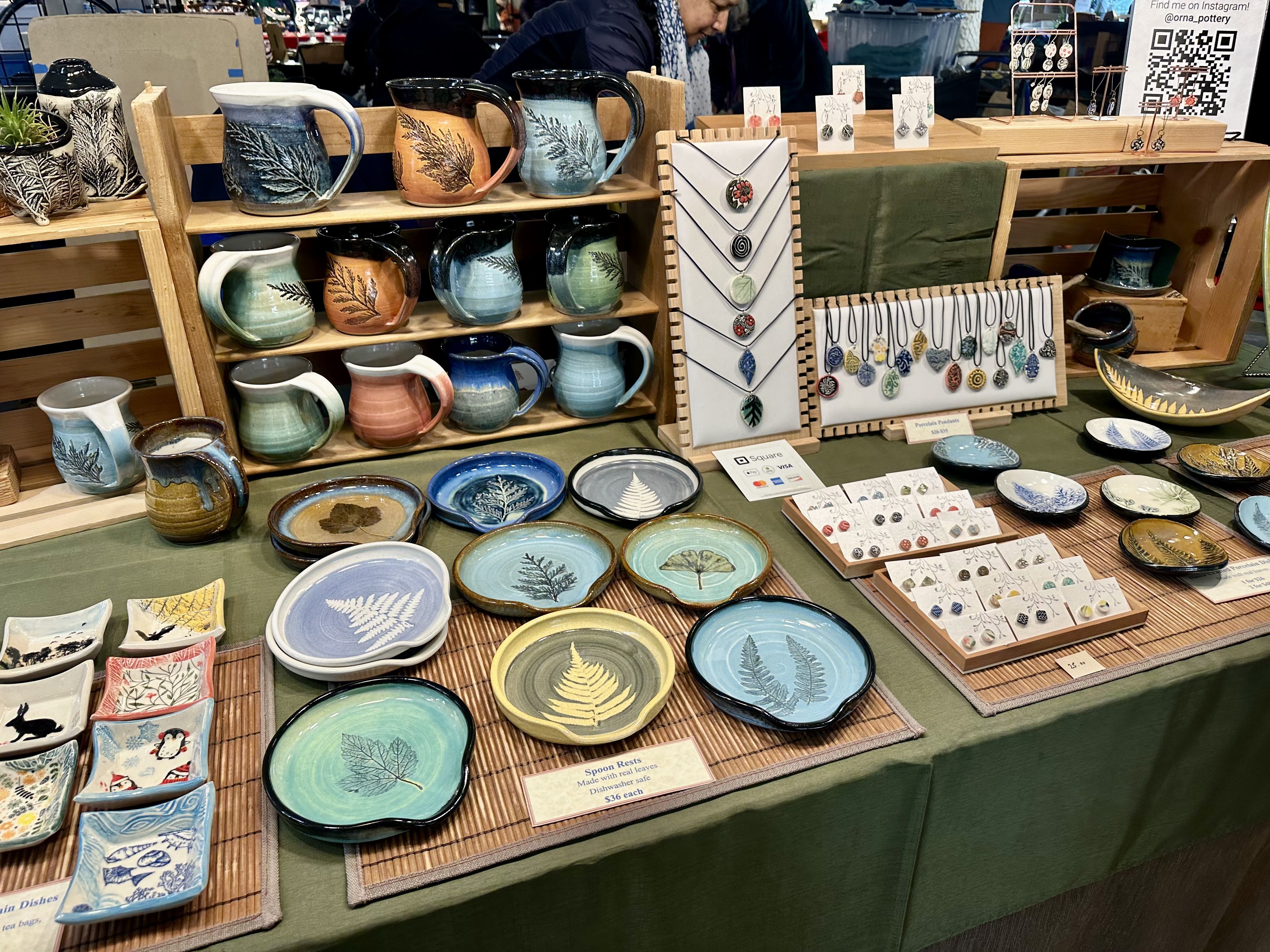 A display of colorful pottery from Orna's Pottery, including pendants, ceramic plates, mugs and more imprinted with leaves and other plant designs.