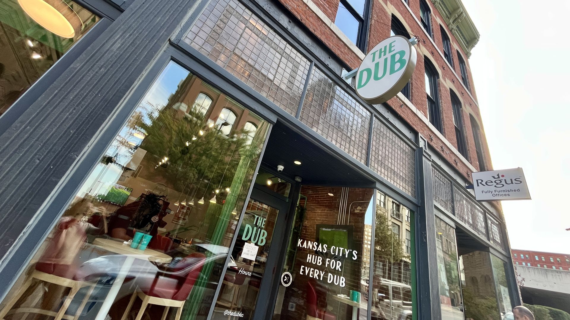 Street view of "The Dub" women's sports bar with green signage on black-framed glass windows and brick building exterior in Kansas City. Reflections of nearby buildings are visible on the glass.