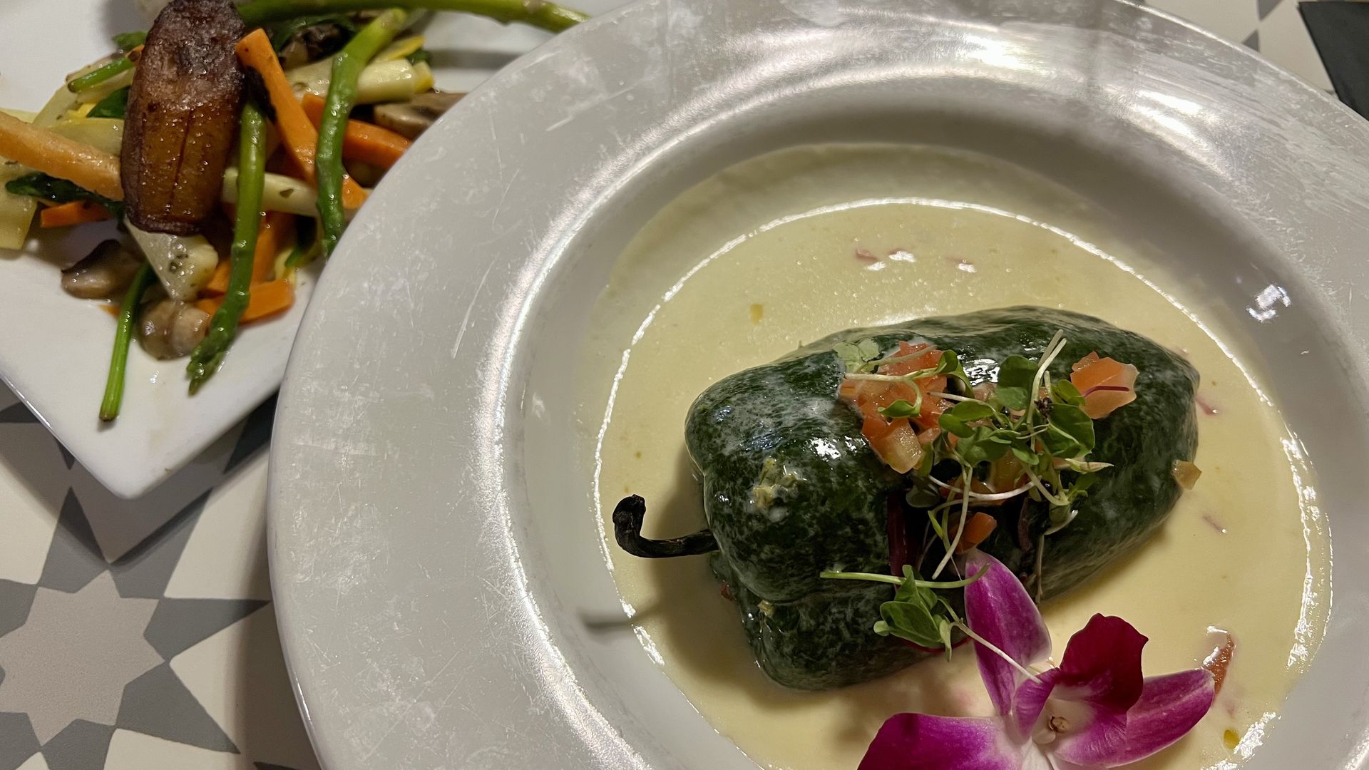 Plate with green stuffed pepper in creamy sauce garnished with microgreens and purple orchid, side plate with rice, sautéed vegetables, and fried plantain on patterned table.